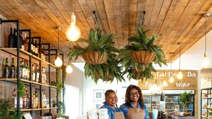 Uma área de bar com prateleiras de madeira repletas de garrafas, plantas penduradas e um letreiro indicando bebidas geladas, sucos de frutas e água.