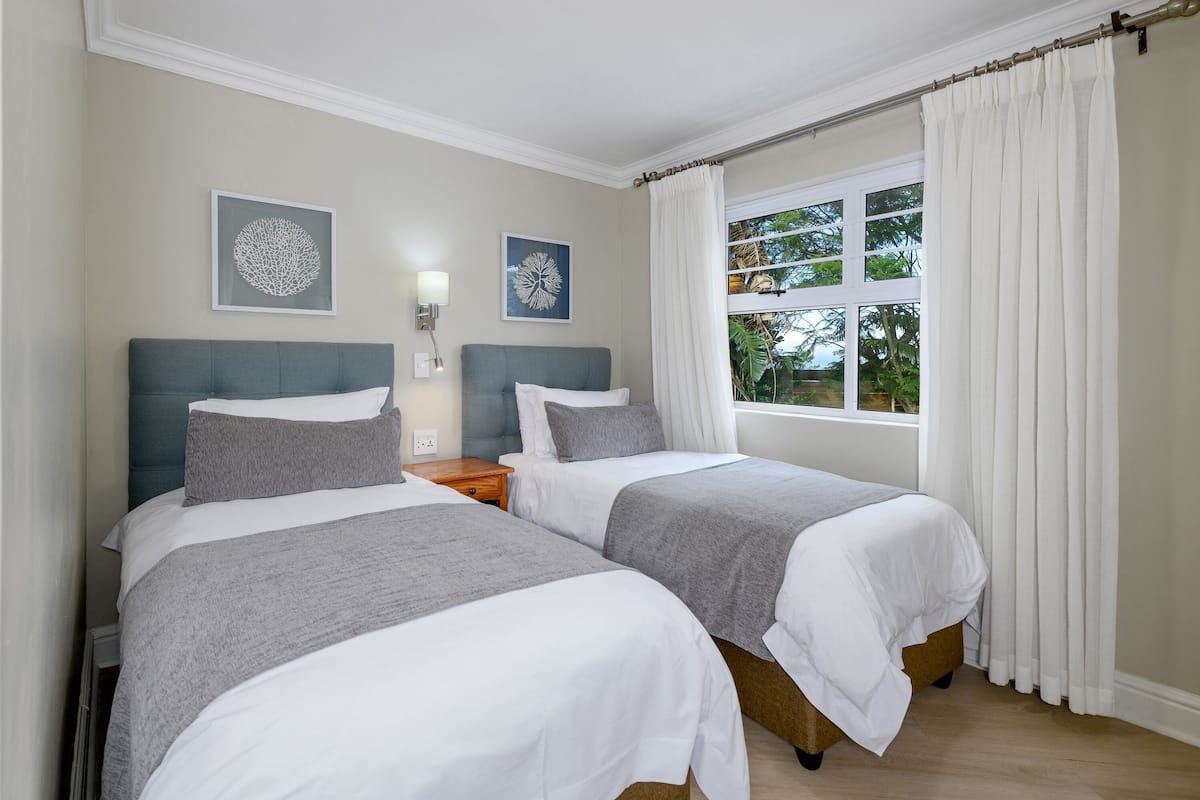 A bedroom with two beds, a nightstand, a wall-mounted lamp, and a window with curtains.
