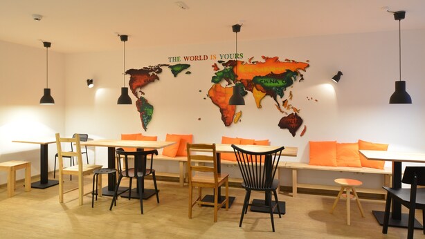 A modern dining area with wooden tables and chairs, orange cushions, and a world map on the wall. - Townside Hostel Bremen (Bremen)