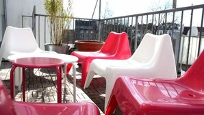 A balcony with red and white plastic chairs and a small table, a potted plant, and a view of buildings in the background. - Townside Hostel Bremen (Bremen)