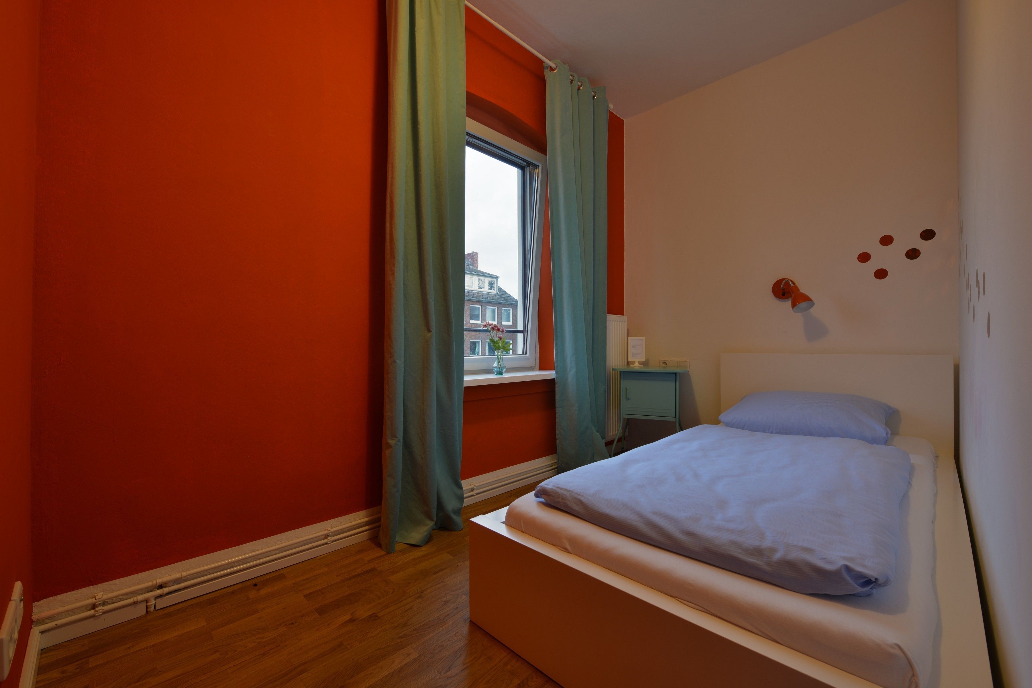 Ein Schlafzimmer mit einem Einzelbett, einem Fenster mit Vorhängen, einem Nachttisch und Blick auf ein Gebäude.
