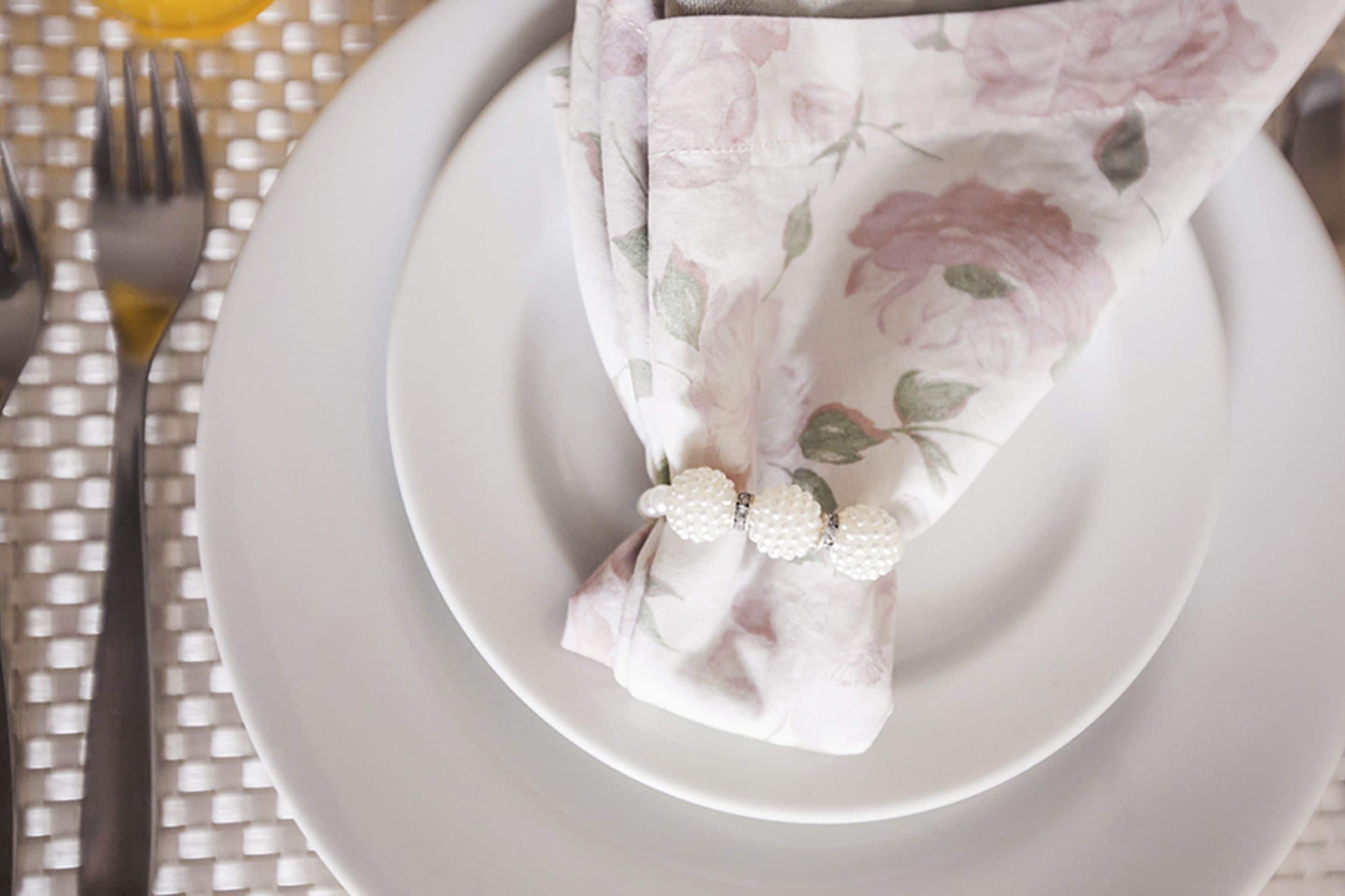A table setting with a white plate, a napkin with a floral pattern, and a pearl accessory.