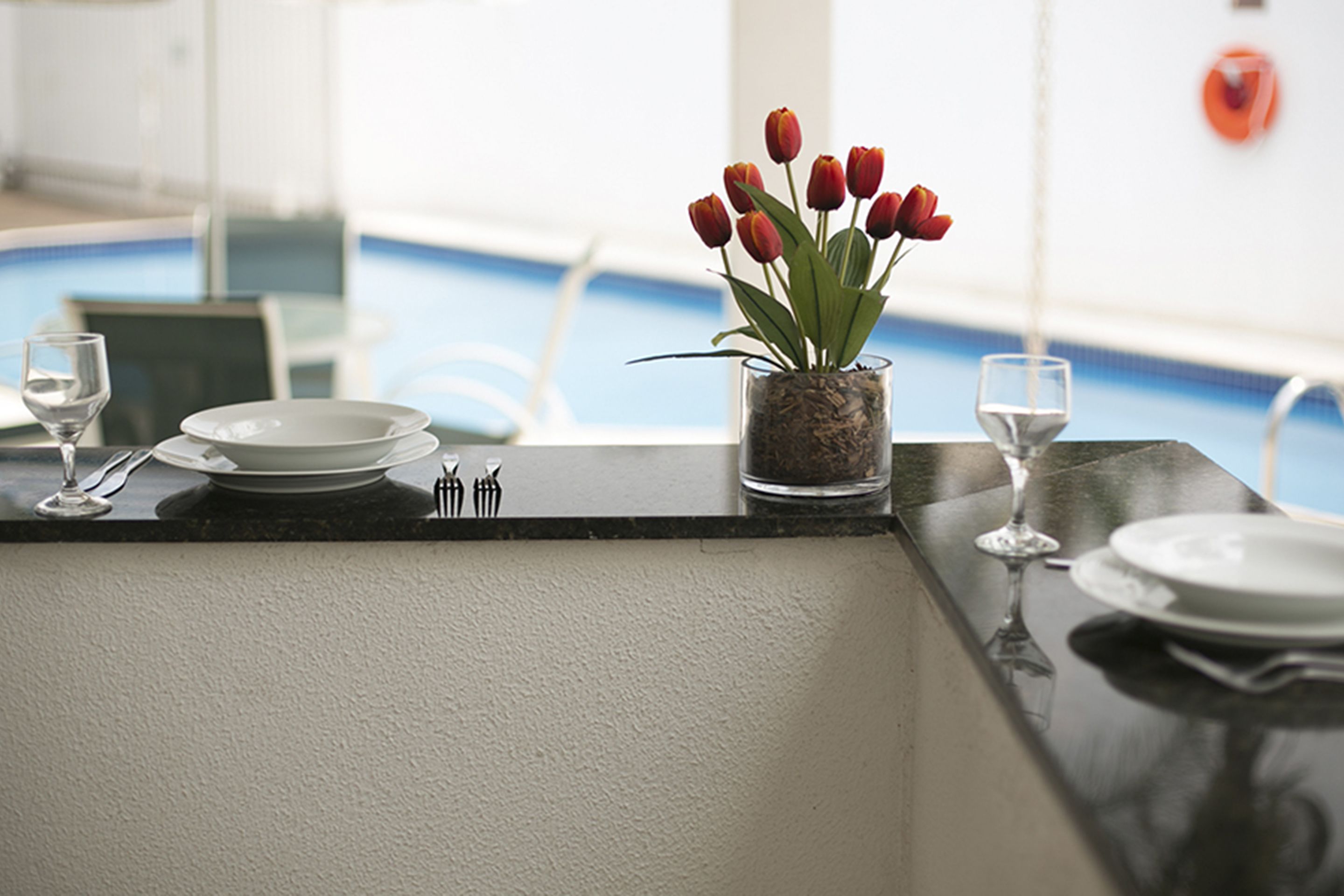 Zona pranzo con vista piscina, un vaso di vetro con tulipani rossi e un tavolo apparecchiato con piatti e posate.