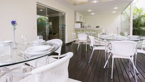 An outdoor dining area with round glass tables and white wicker chairs, connected to a kitchen area with white cabinets and a view of greenery. - Verona Hplus Long Stay (Lago Norte)