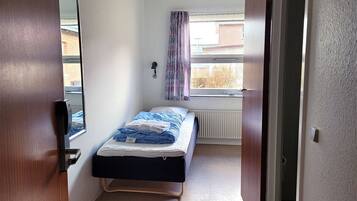 Una piccola camera con un letto singolo, una finestra, un termosifone e una porta.