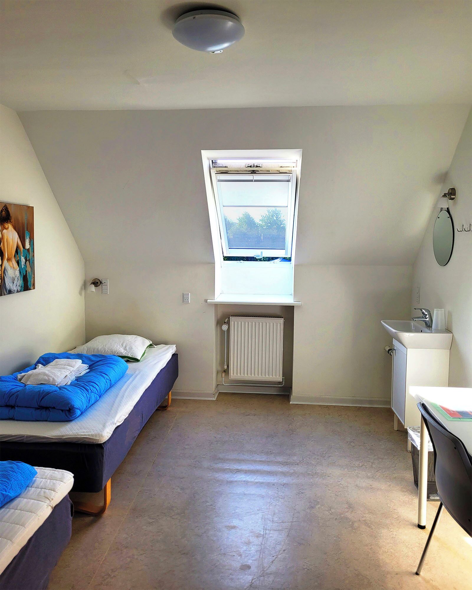 Una habitación pequeña y bien iluminada, con dos camas, una ventana, un radiador, un lavabo y un espejo.