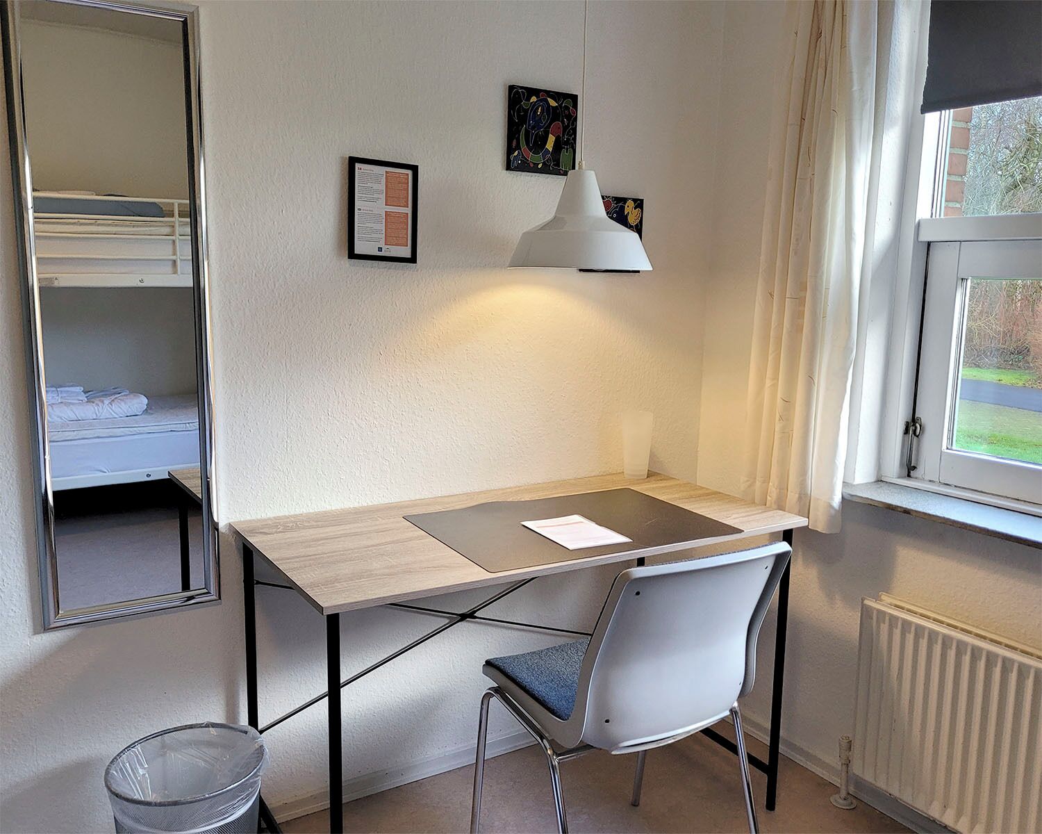 Een kleine hotelkamer met een houten bureau, een witte stoel, een spiegel en een raam met gordijnen.