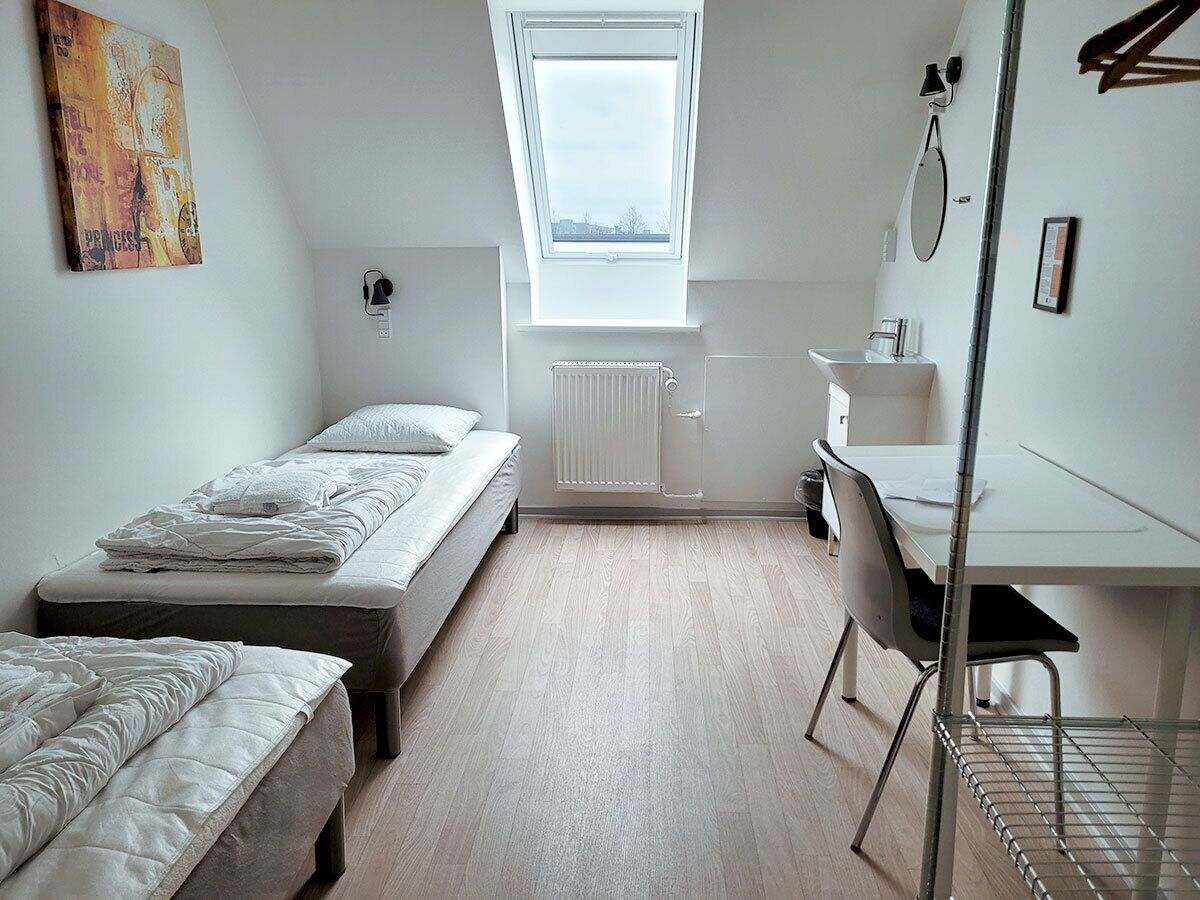 Quarto de estudantes com duas camas, uma escrivaninha, um radiador e um clarabóia.