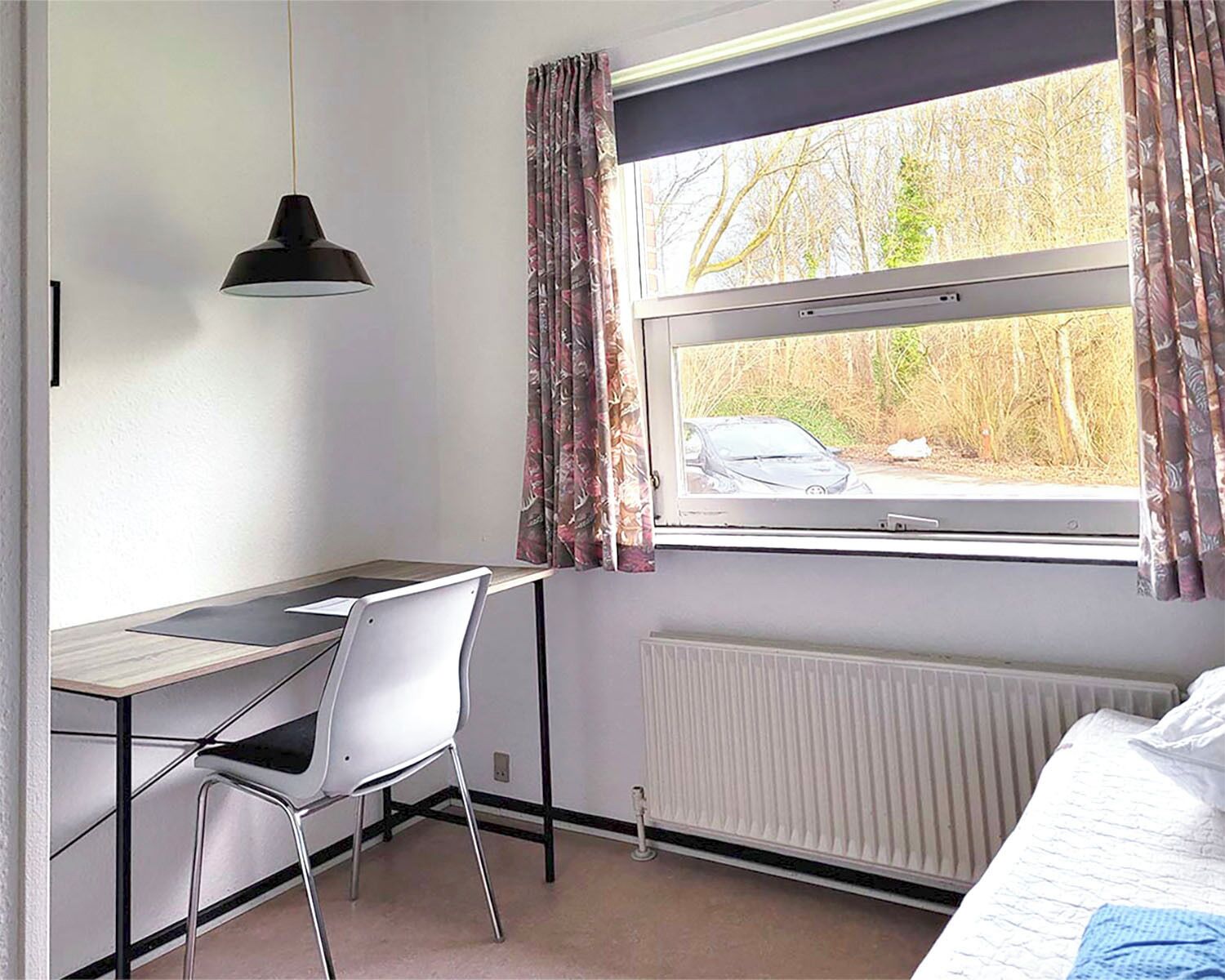A room with a desk, chair, radiator, and a window with curtains.
