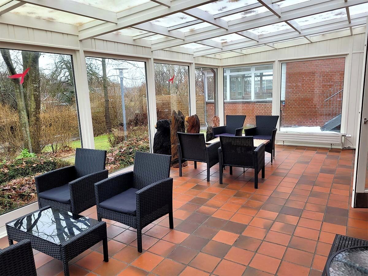 Un patio couvert, aménagé avec des meubles en osier et une table en verre, donnant sur un mur de briques et un jardin.