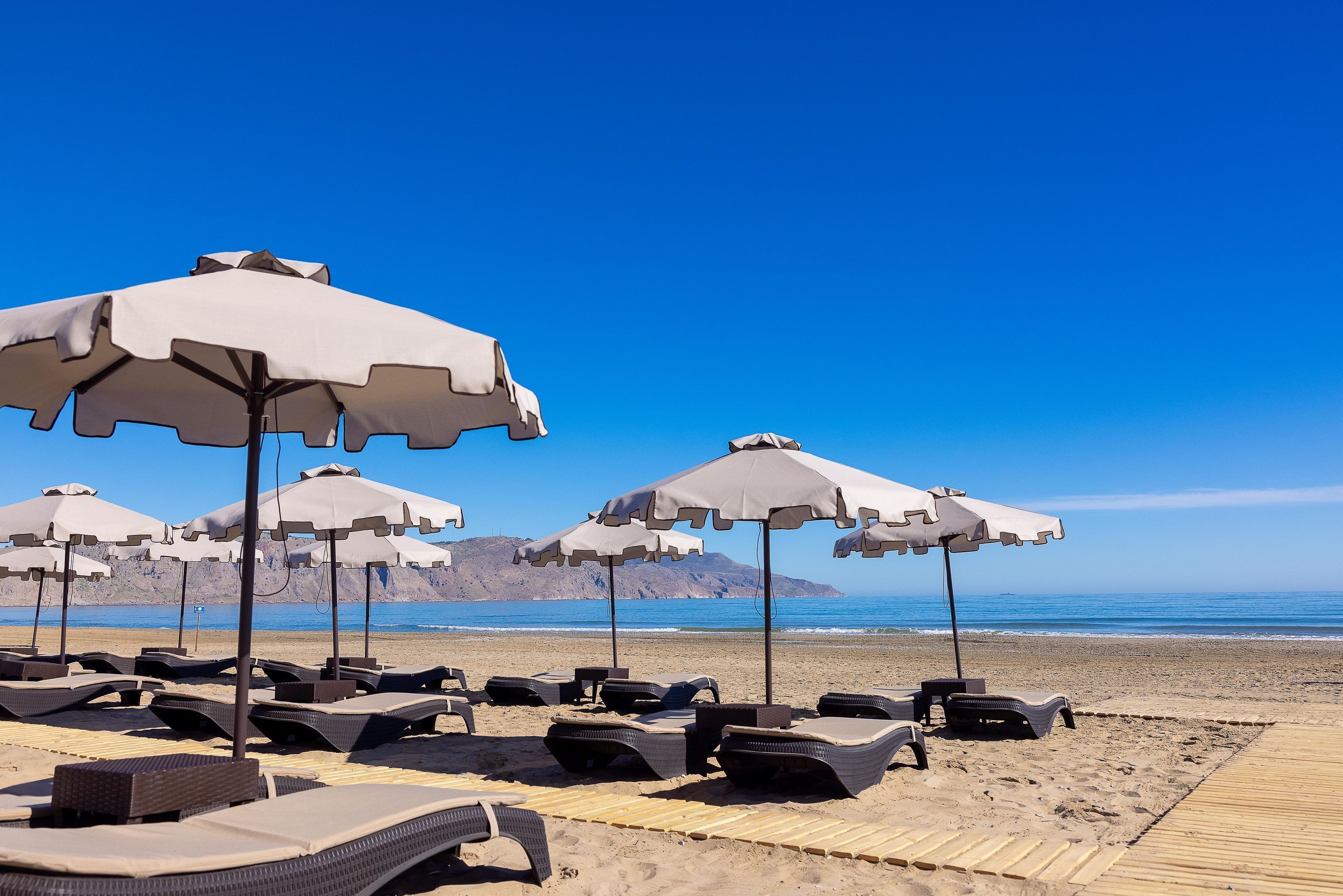 Beach umbrellas, lounge chairs, and a wooden boardwalk on a sandy beach with a clear sky and mountains in the background.