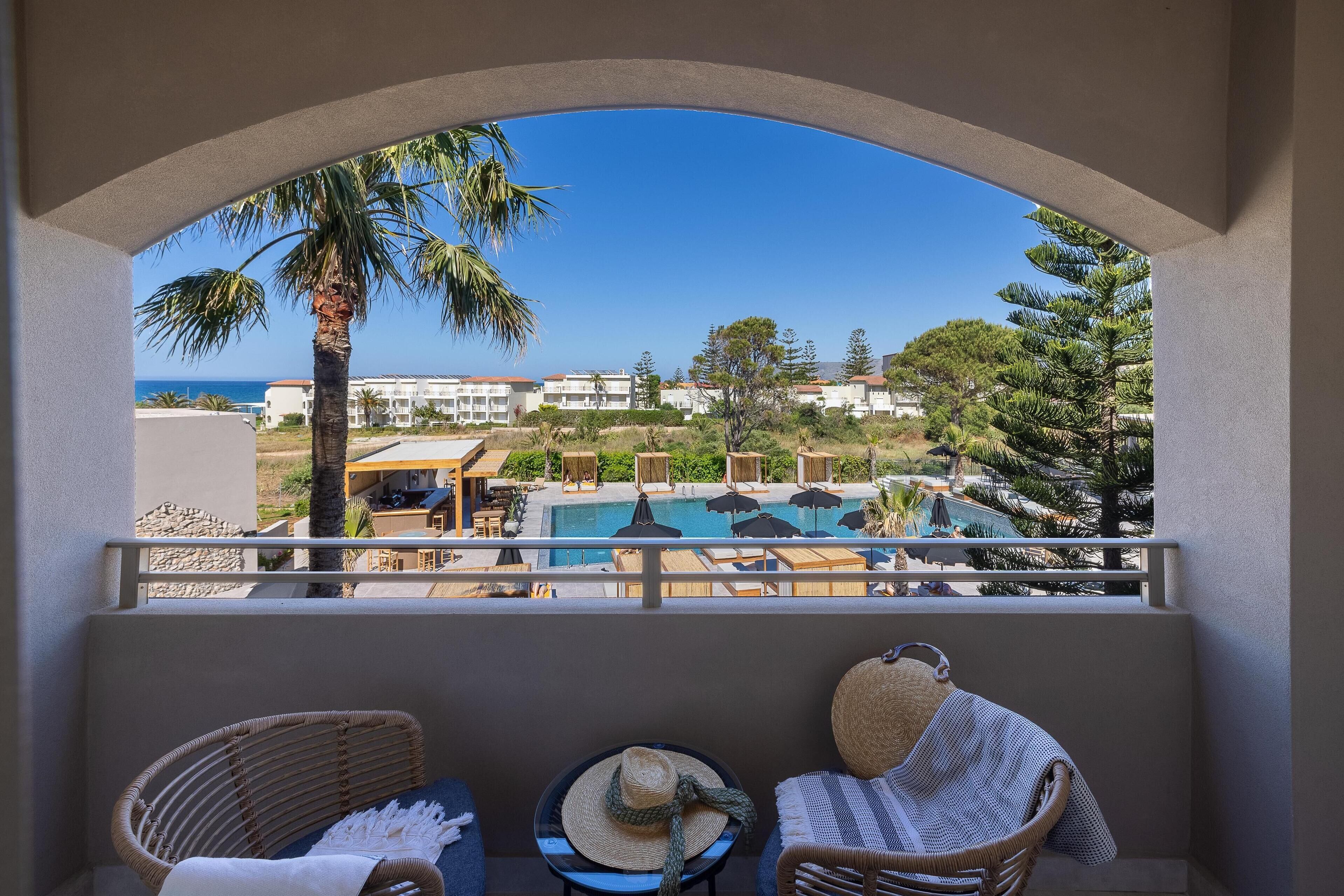 A balcony with wicker chairs, a small table, and a view of a pool and buildings.