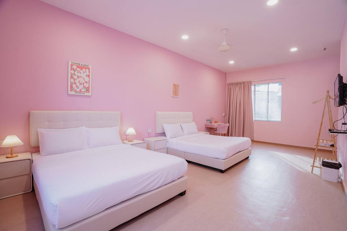 A pink-themed hotel room with two beds, a bedside table with a lamp, a small table with a chair, a window with curtains, and a ladder against the wall.