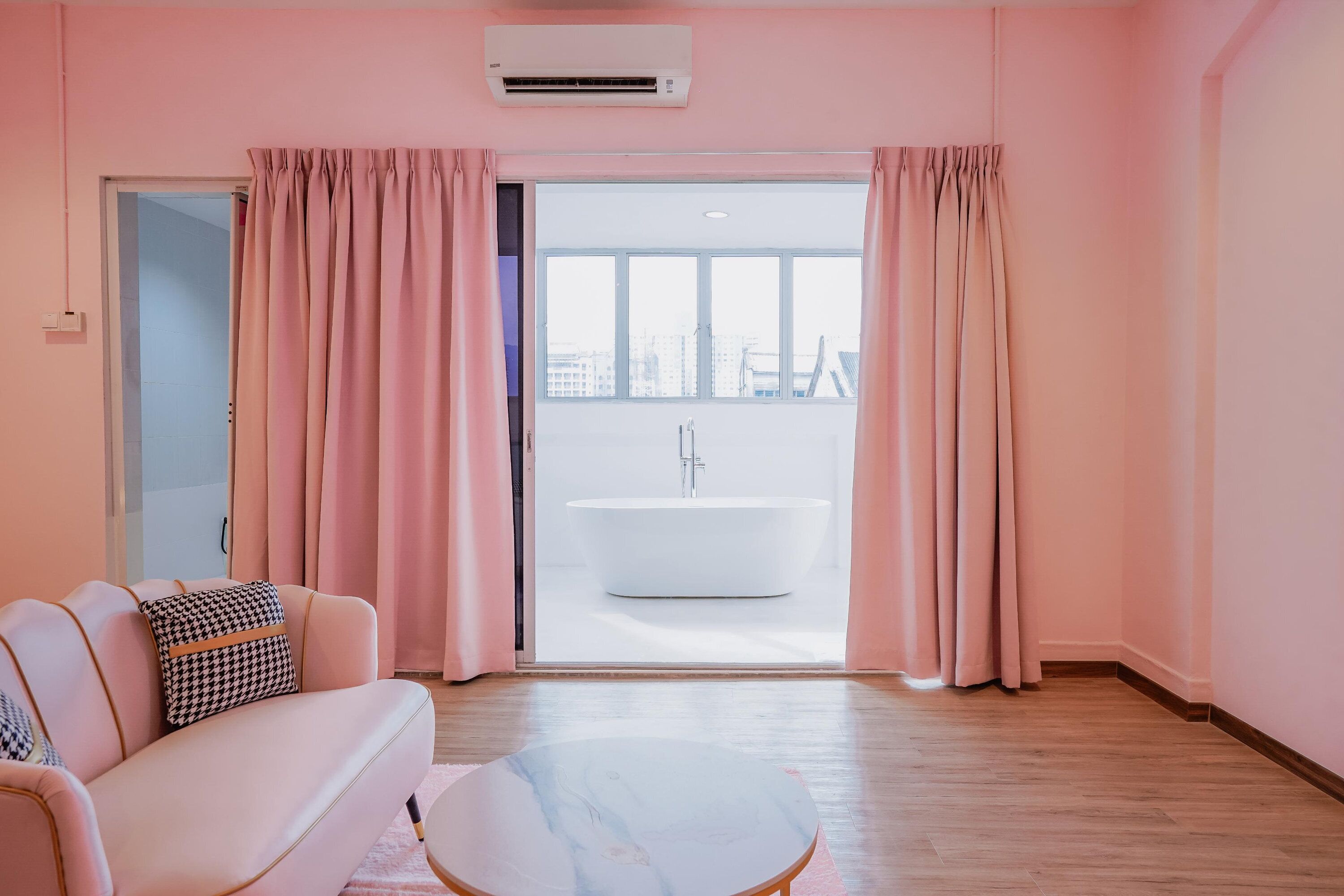 A pink-themed room with a sofa, a marble coffee table, and a view of a white bathtub through pink curtains.
