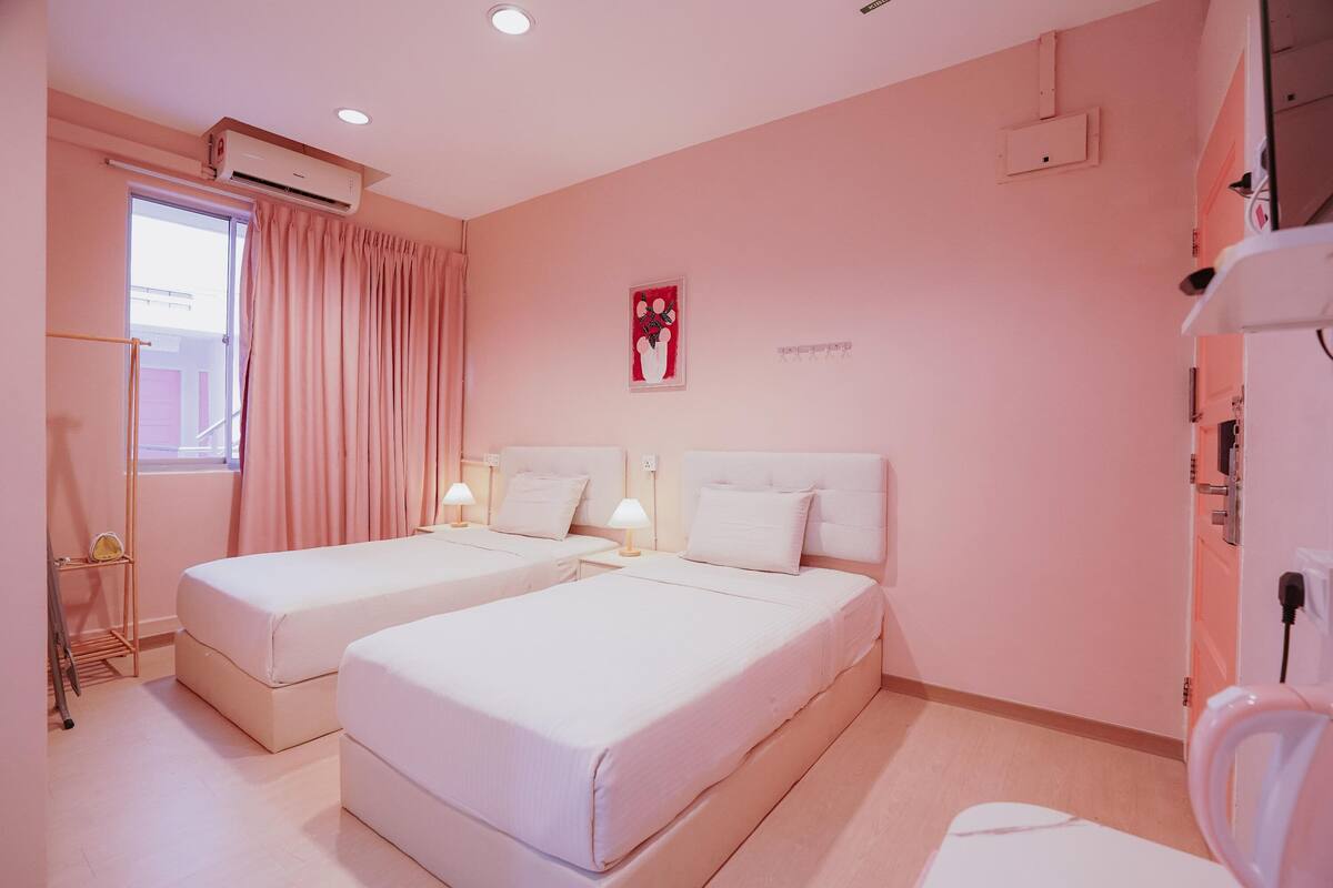 A hotel room with two beds, a window with curtains, an air conditioning unit, a bedside table with a lamp, and a wall-mounted TV.