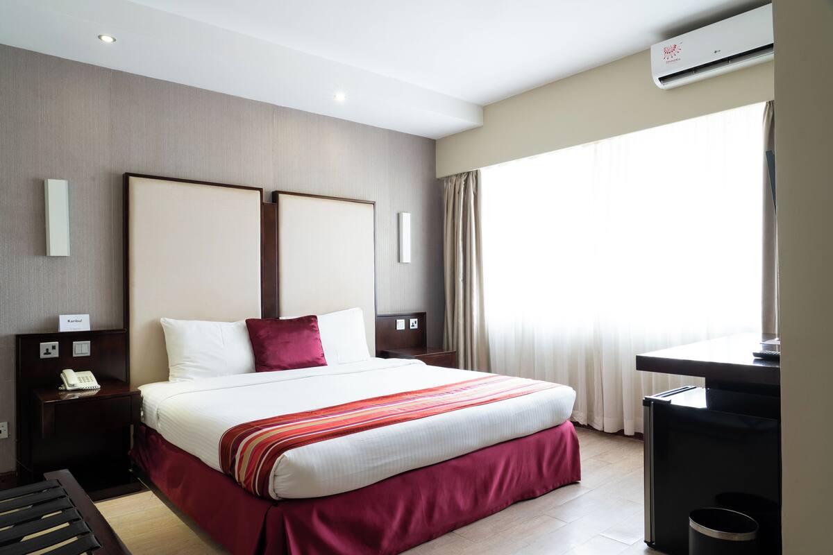 A hotel room with a large bed, a desk, a trash bin, and an air conditioning unit.