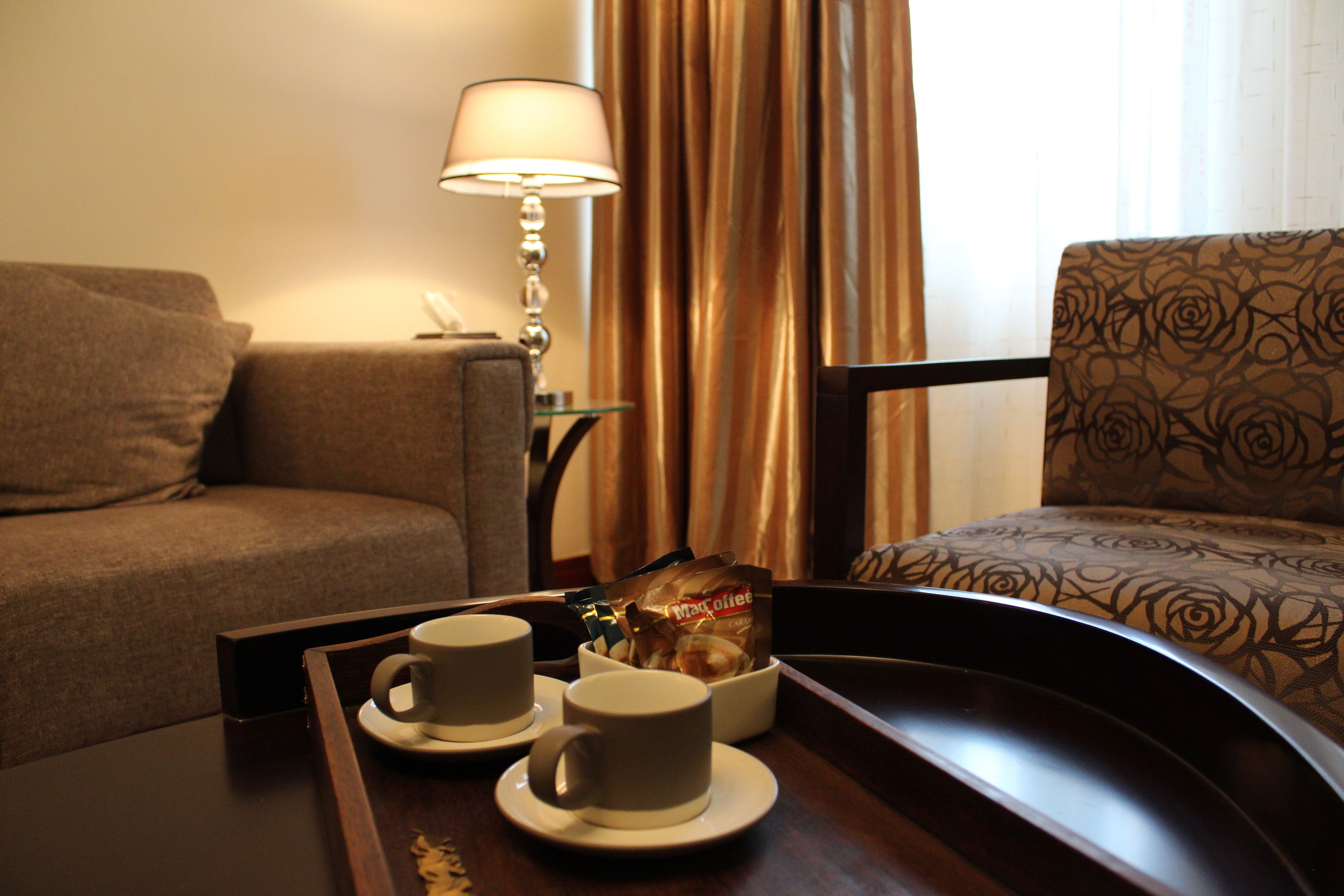 A hotel room with a sofa, a chair, a tray with two cups and a coffee packet, and a lamp.