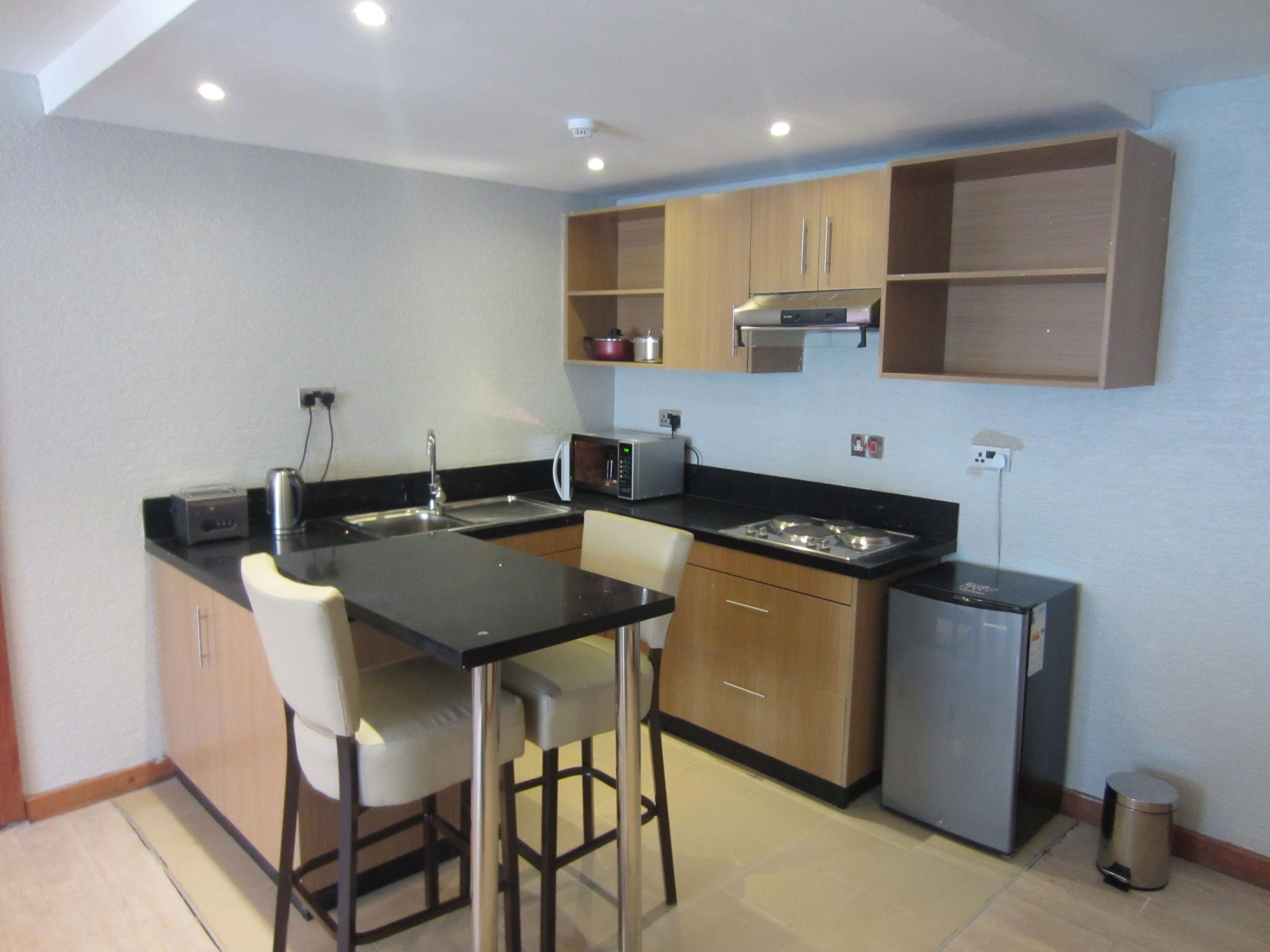 A compact kitchen with a black countertop, white bar stools, wooden cabinets, a stainless steel refrigerator, and a microwave.