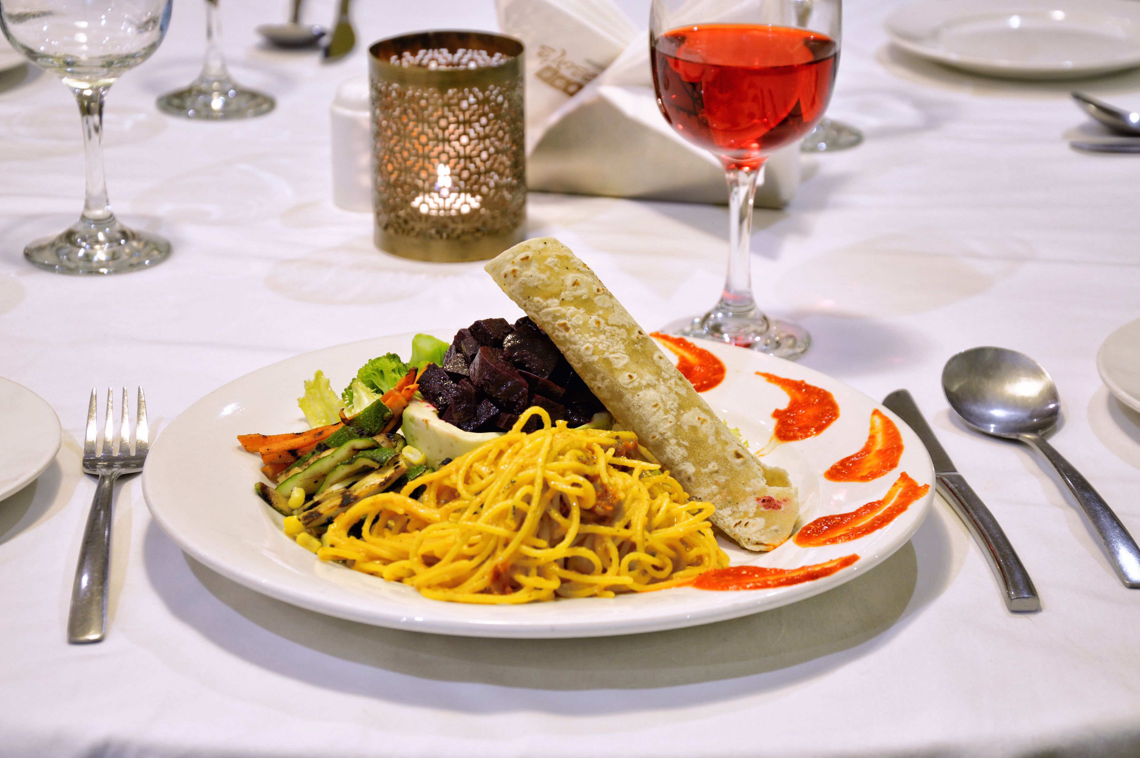 A plate of spaghetti with vegetables and a breadstick, accompanied by a glass of red wine, on a white tablecloth.
