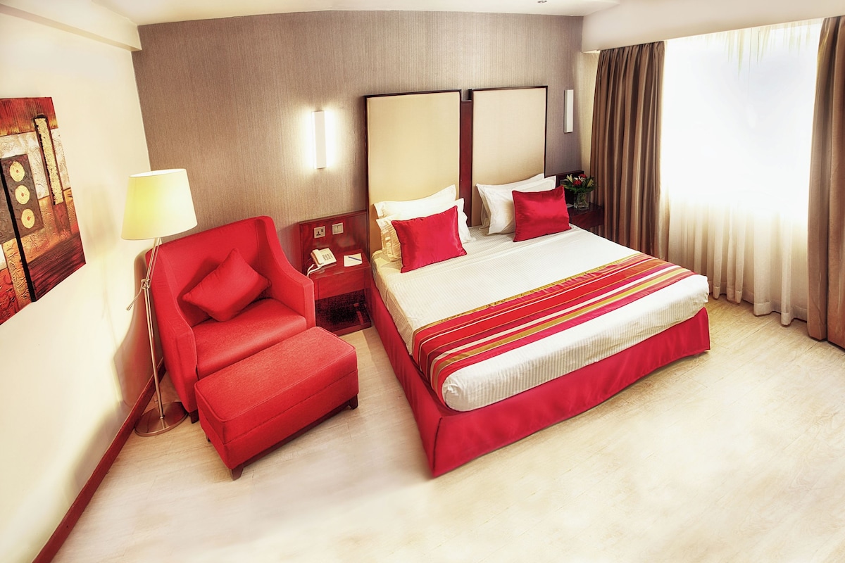 Una habitación de hotel con una cama grande, un sillón rojo, una mesita de noche con lámpara y un espejo amplio.
