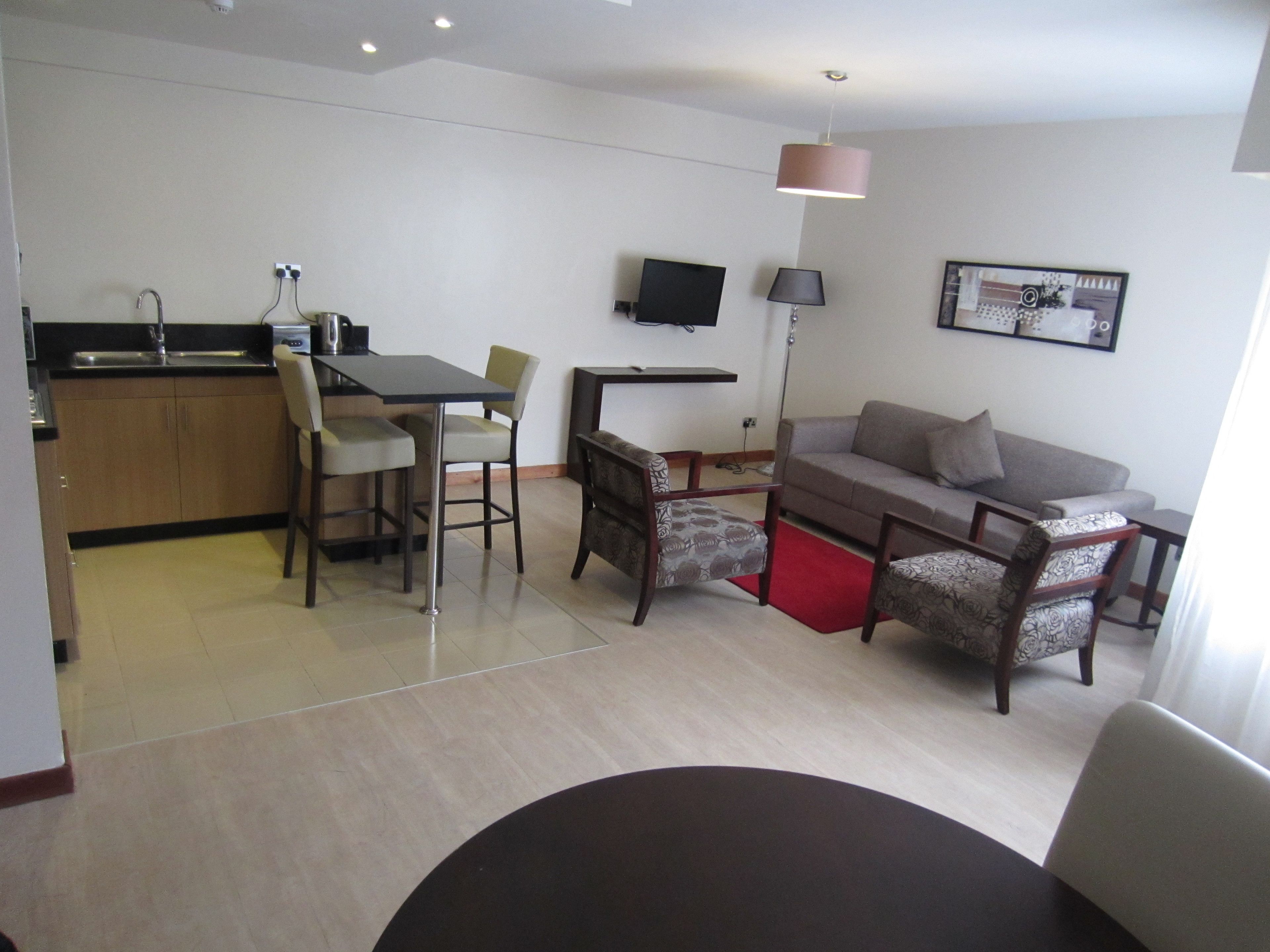 Una sala de estar moderna con un sofá, dos sillones, una mesa de comedor con sillas, una zona de cocina y un televisor de pantalla plana montado en la pared.
