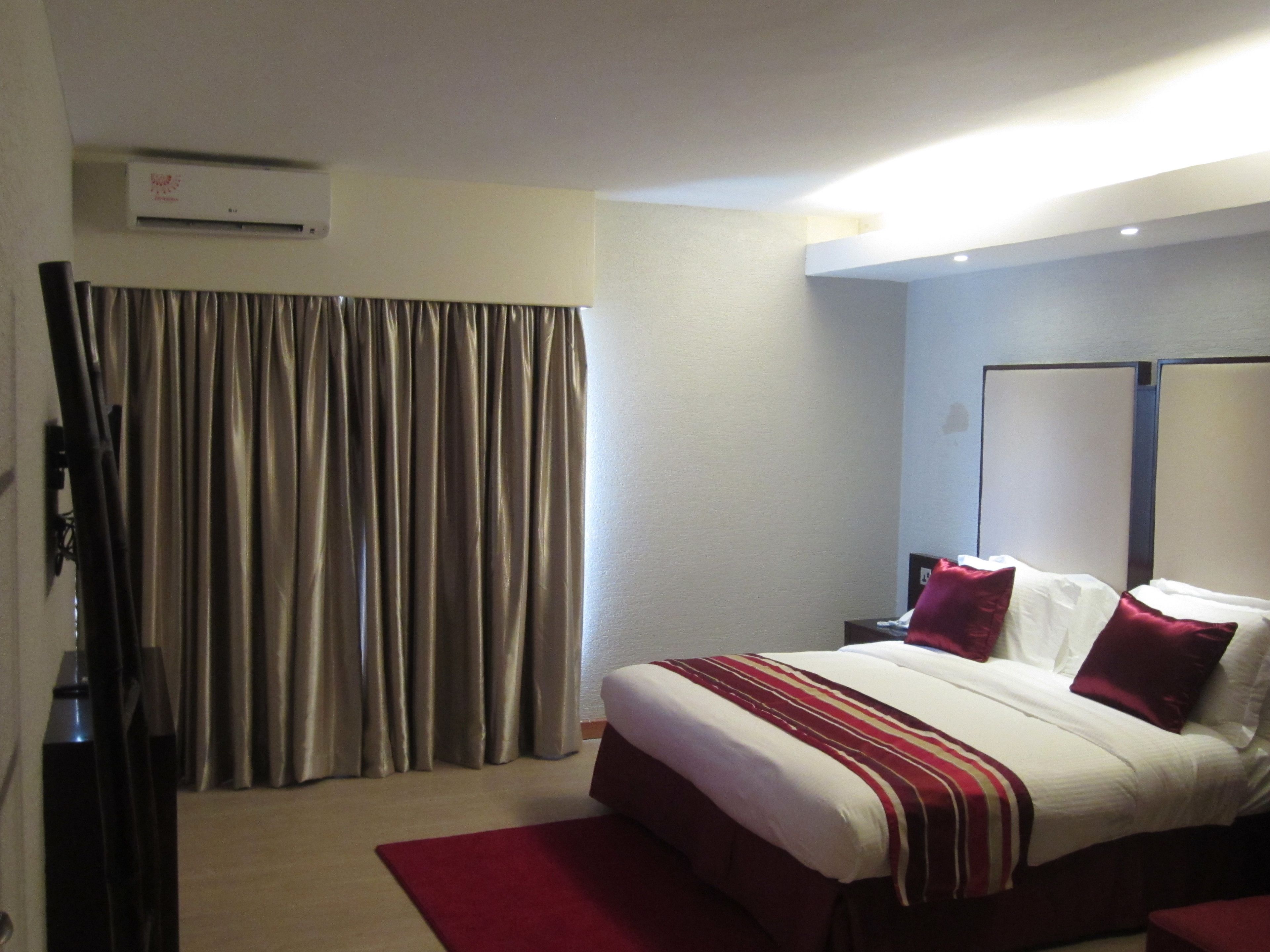 A hotel room with a bed, curtains, a flat-screen TV, and an air conditioning unit.