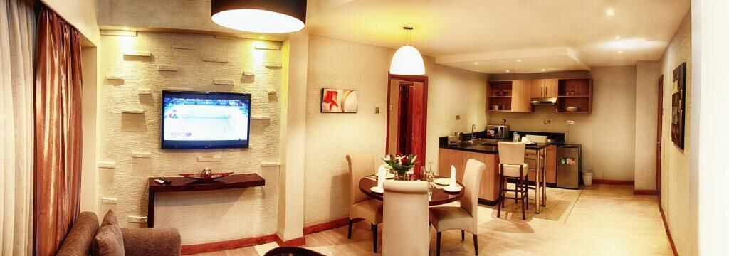 A modern living room with a flat-screen TV mounted on the wall, a dining area with a round table and chairs, and a kitchen area with wooden cabinets and a countertop.