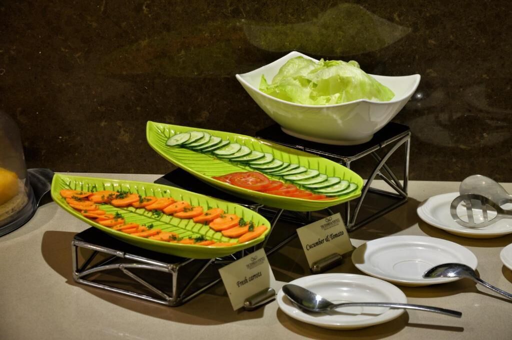A buffet setup with labeled dishes of fresh carrots, cucumber and tomato, and lettuce.