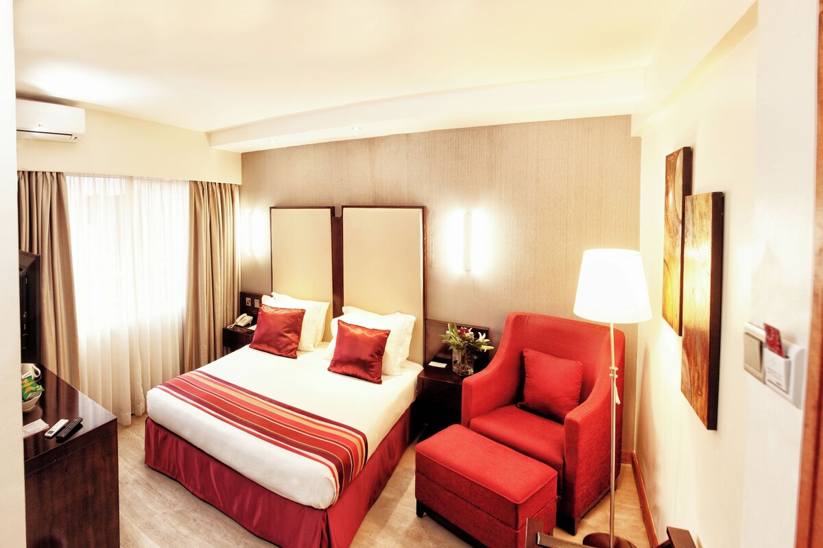 A hotel room with a large bed, a red armchair, a desk with a lamp, and a trash bin.