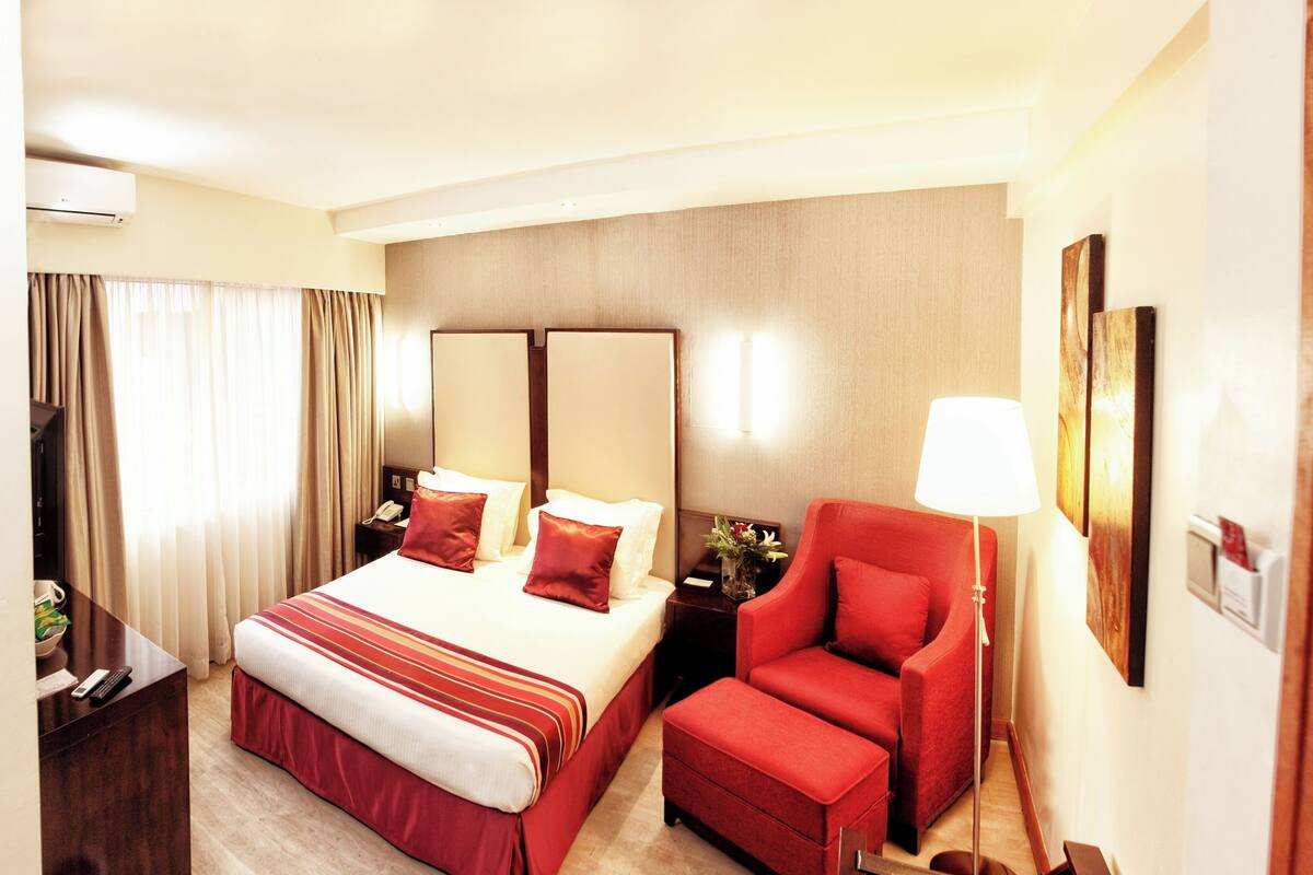 A hotel room with a large bed, a red armchair, a small table, and a lamp.