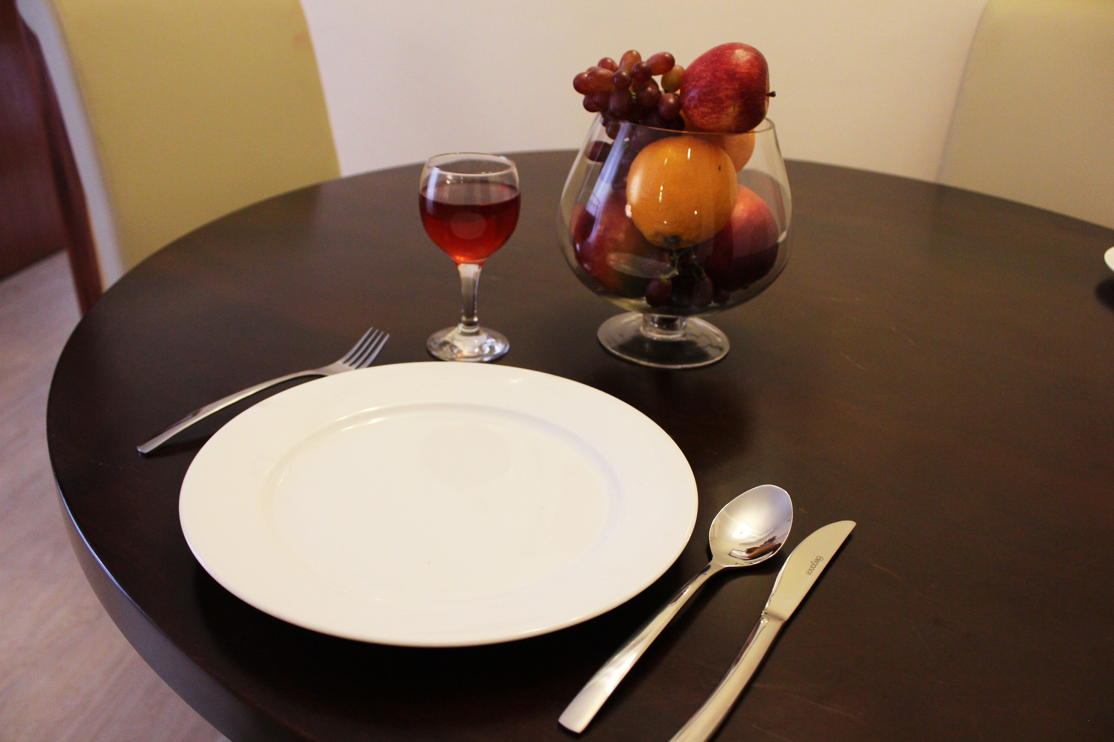 A dining table with a white plate, a glass of red wine, and a fruit bowl containing grapes, apples, and an orange.