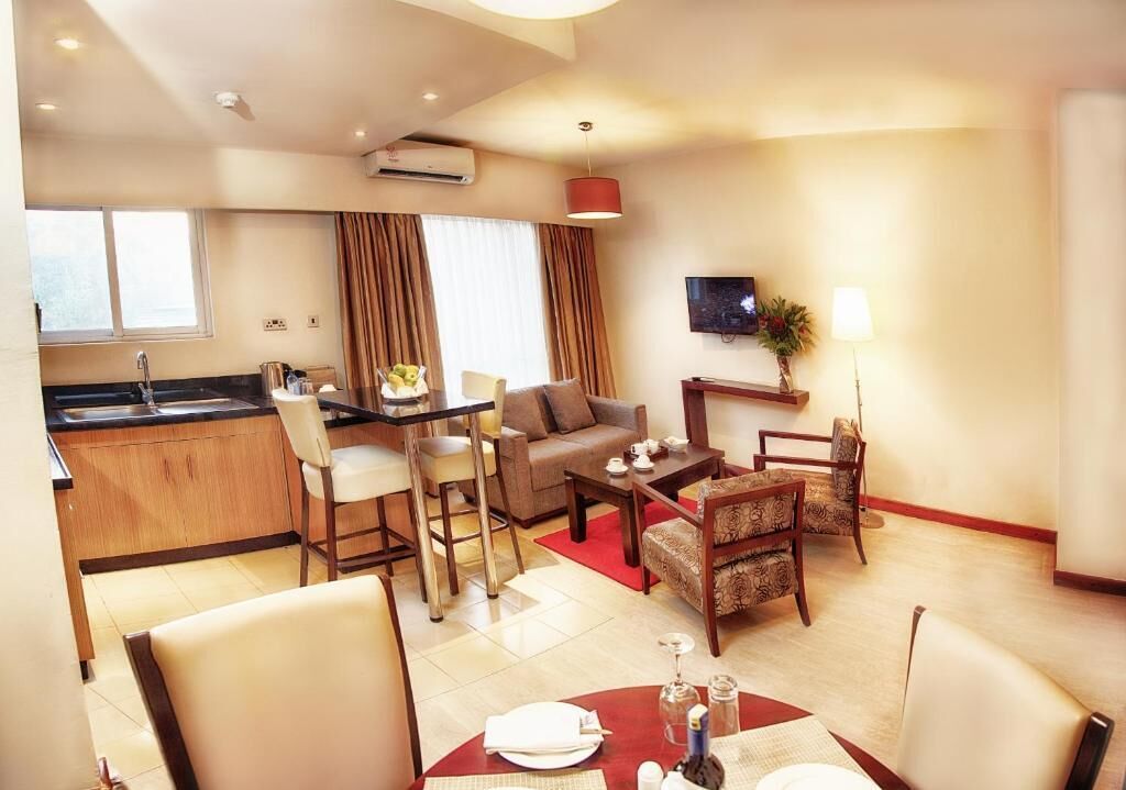 A modern hotel room with a kitchenette, a sofa, a dining table, and a TV.