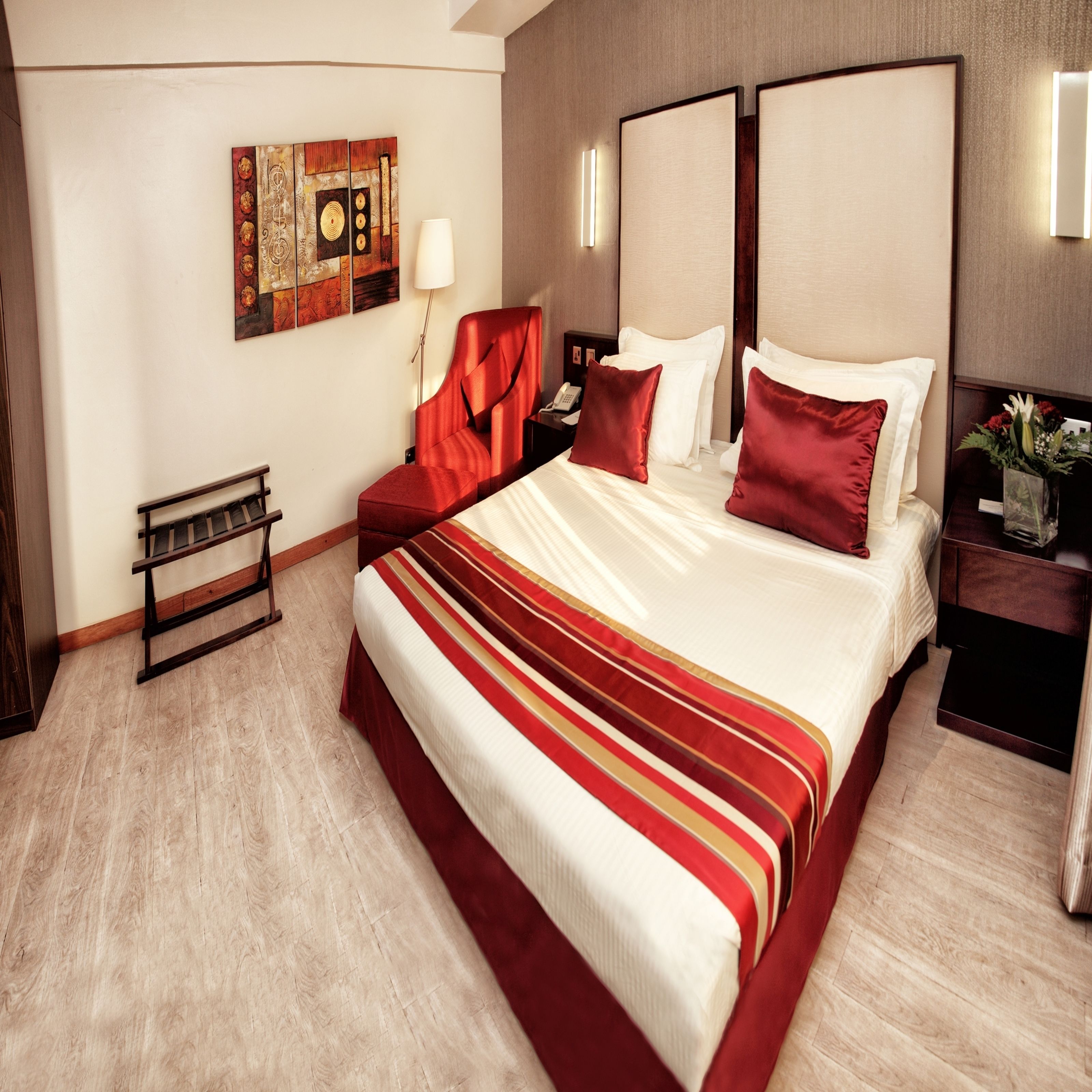 Habitación de hotel con una cama grande, una mesita de noche con lámpara, un pequeño banco y una obra de arte colgada en la pared.