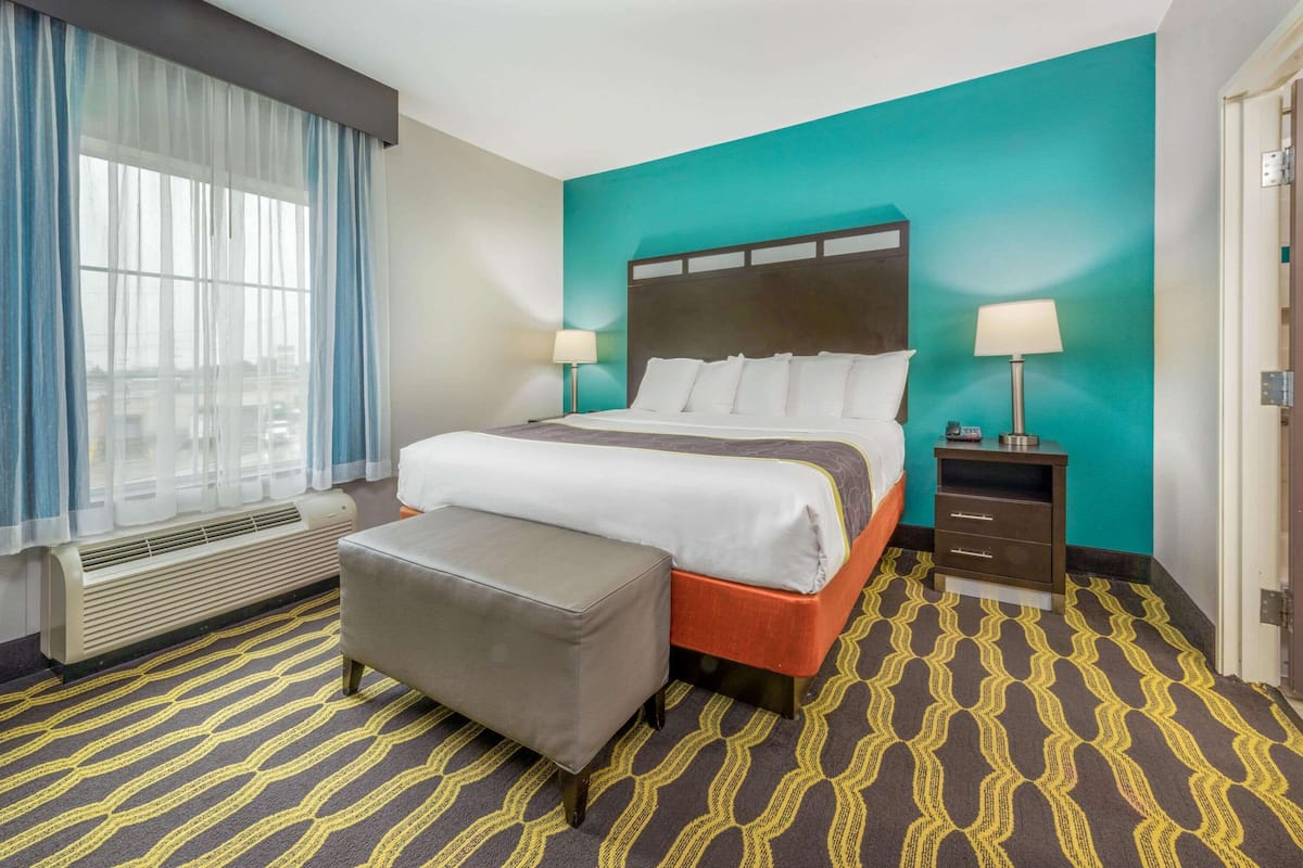 Une chambre d’hôtel comprenant un lit, des tables de chevet, un banc, une fenêtre avec des rideaux, un radiateur et une moquette à motifs.