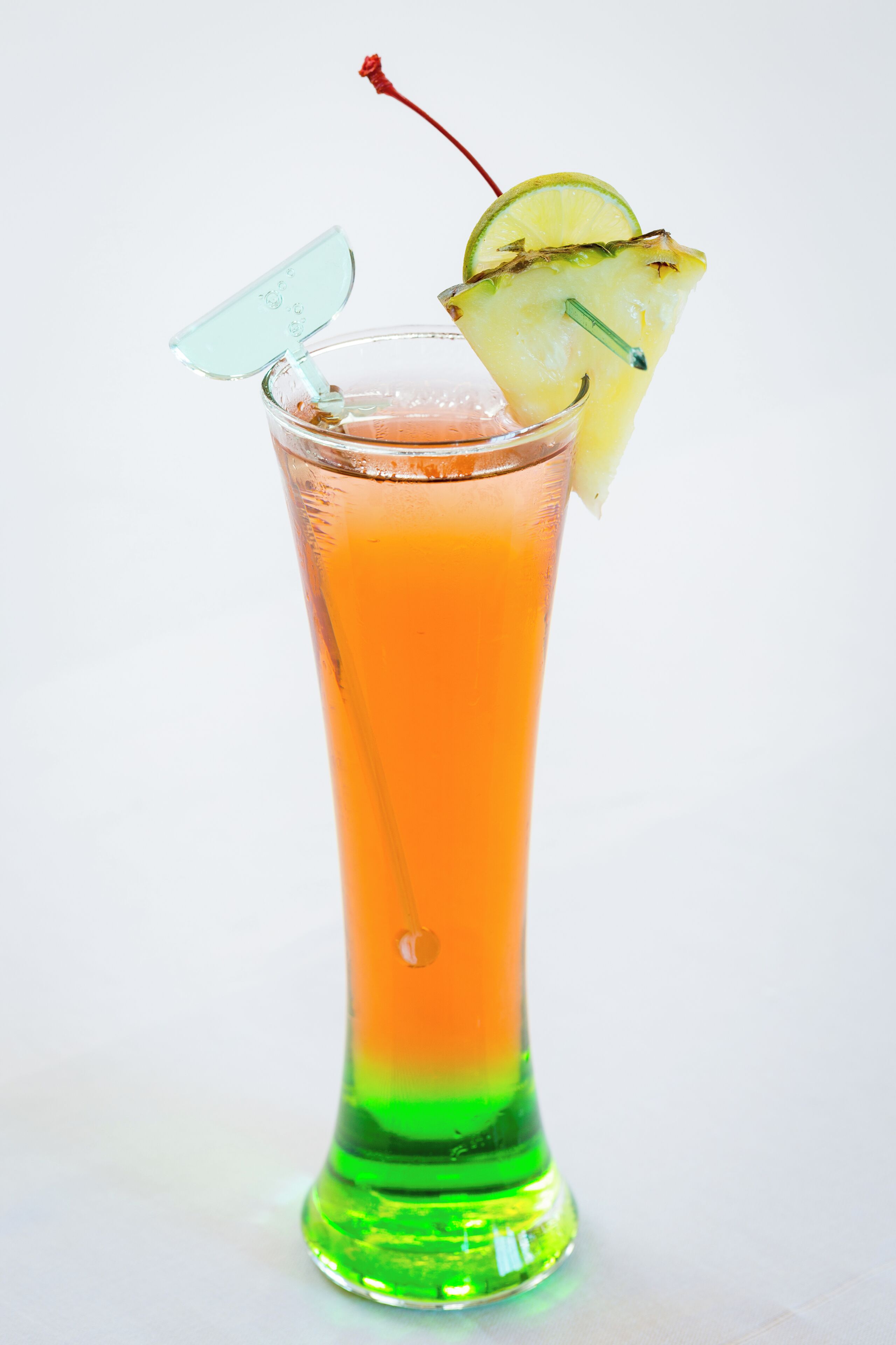 A tall glass with a gradient from green at the bottom to orange at the top, containing a drink with a lime slice and a cherry on the rim.