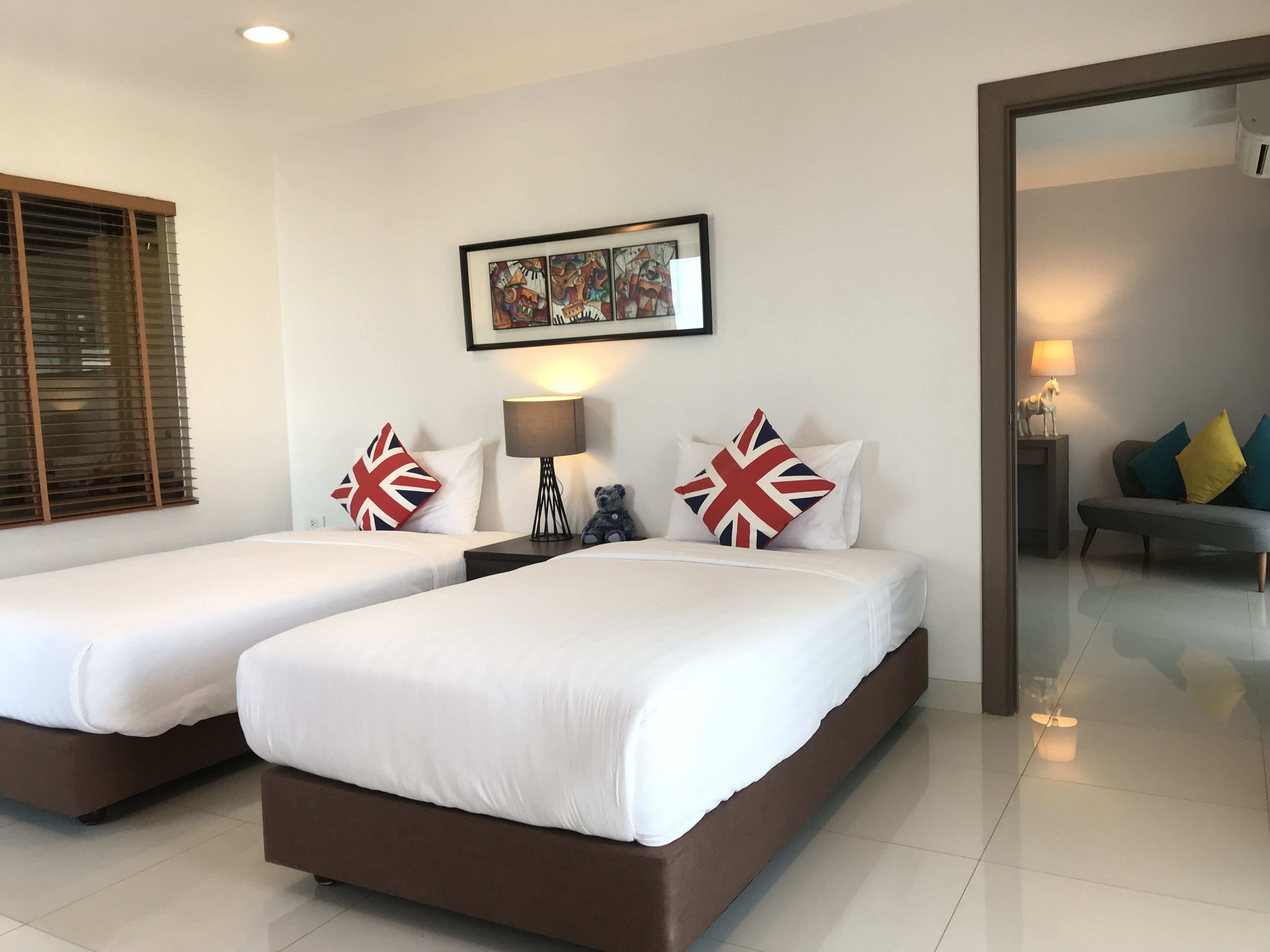 A hotel room with two beds, a framed artwork above the beds, a lamp on the nightstand, and a sofa with cushions in the corner.