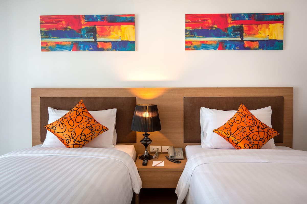 Une chambre d’hôtel avec deux lits, chacun agrémenté de draps blancs et d’oreillers à motifs orange. Une table de chevet se trouve entre les lits, sur laquelle reposent une lampe, un téléphone et un bloc-notes. Au-dessus des lits, deux tableaux abstraits sont accrochés au mur.