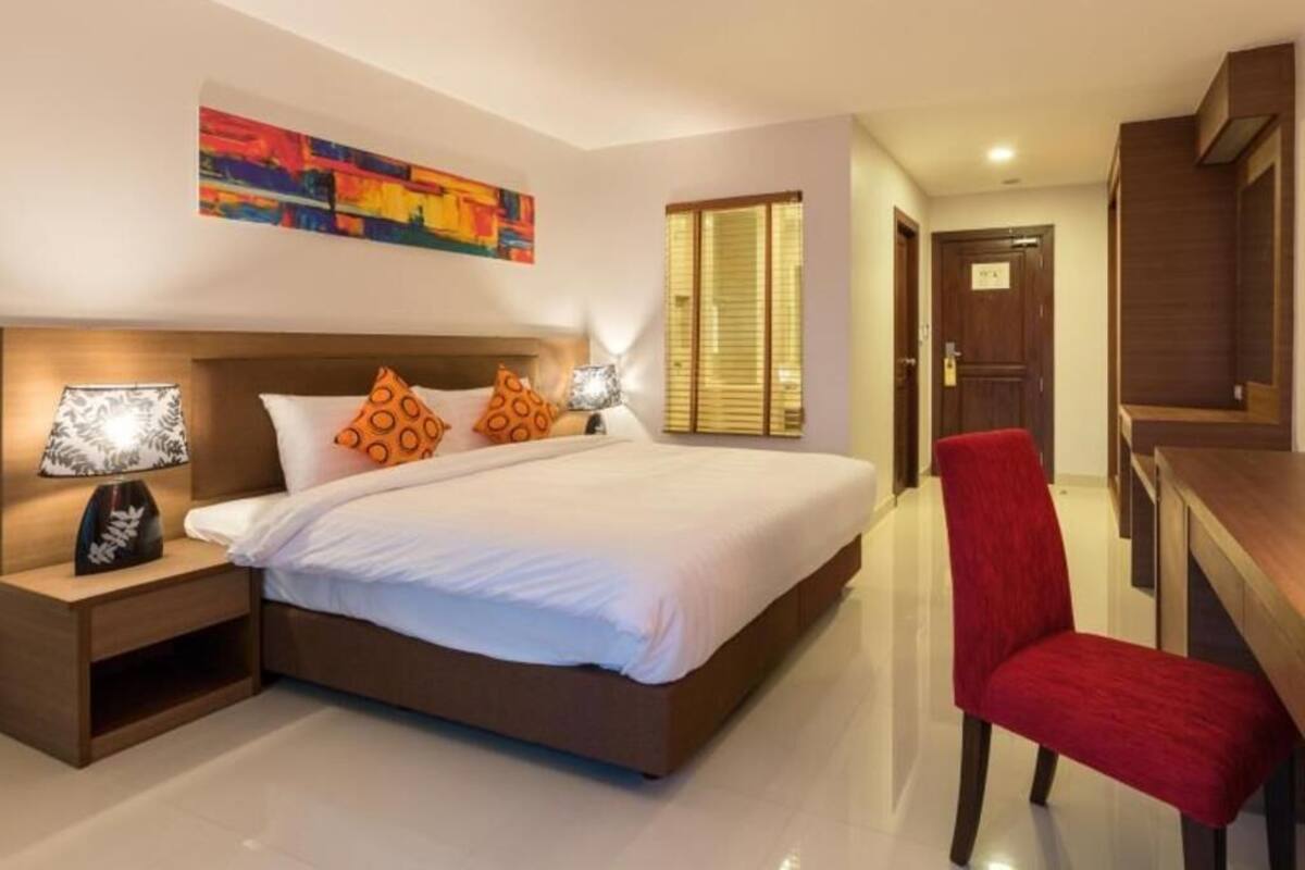 Une chambre d’hôtel avec un grand lit, des lampes de chevet, un fauteuil rouge, un bureau en bois et un tableau abstrait aux couleurs vives accroché au mur.