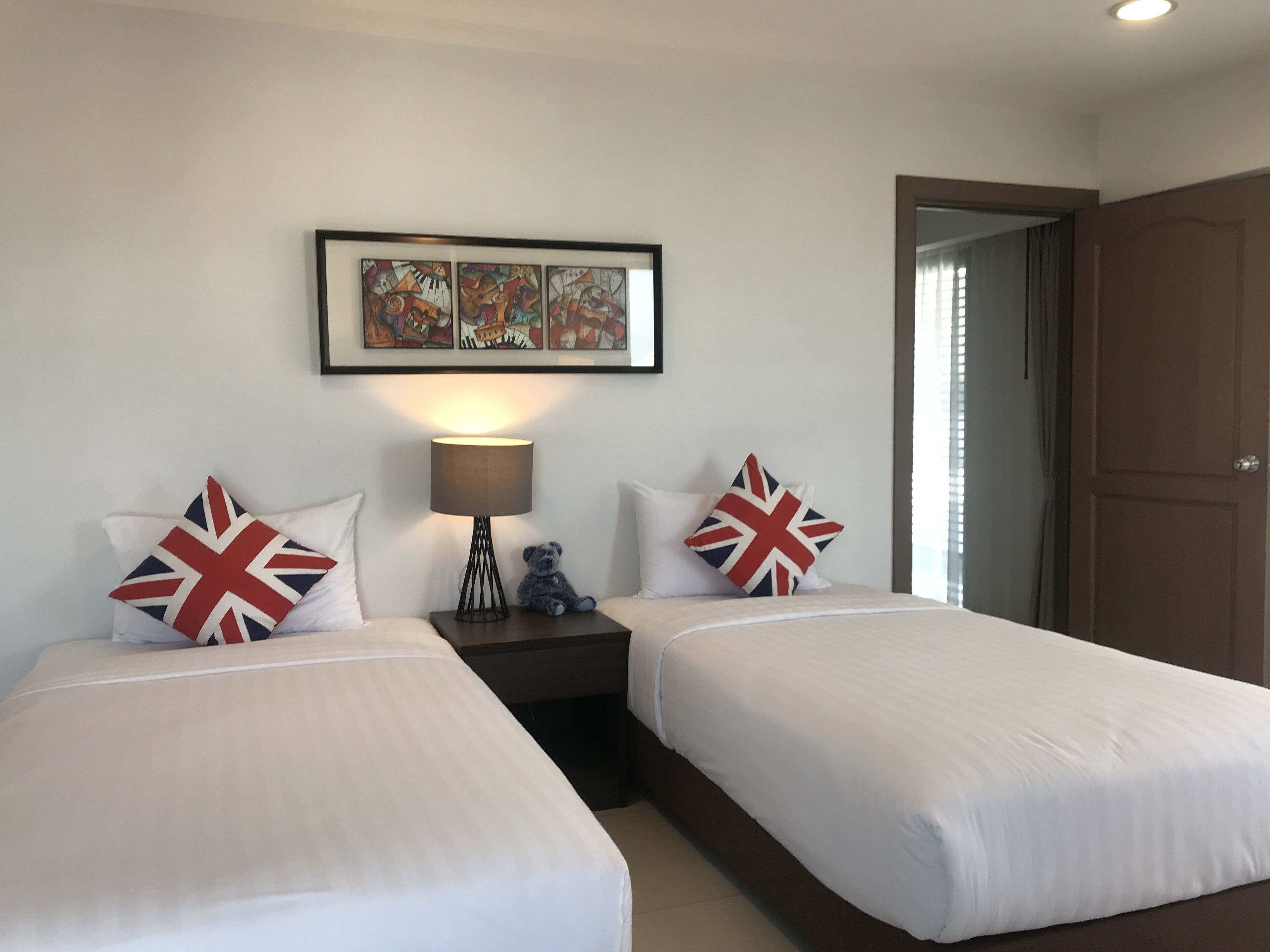 A hotel room with two beds, each with a pillow featuring the Union Jack design. There is a bedside table with a lamp and a small teddy bear. Above the beds, there is a framed artwork. The room has a door on the right side.