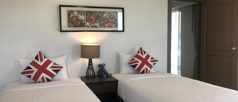 A hotel room with two beds, each with a pillow featuring the Union Jack design. There is a bedside table with a lamp and a small teddy bear. Above the beds, there is a framed artwork. The room has a door on the right side.