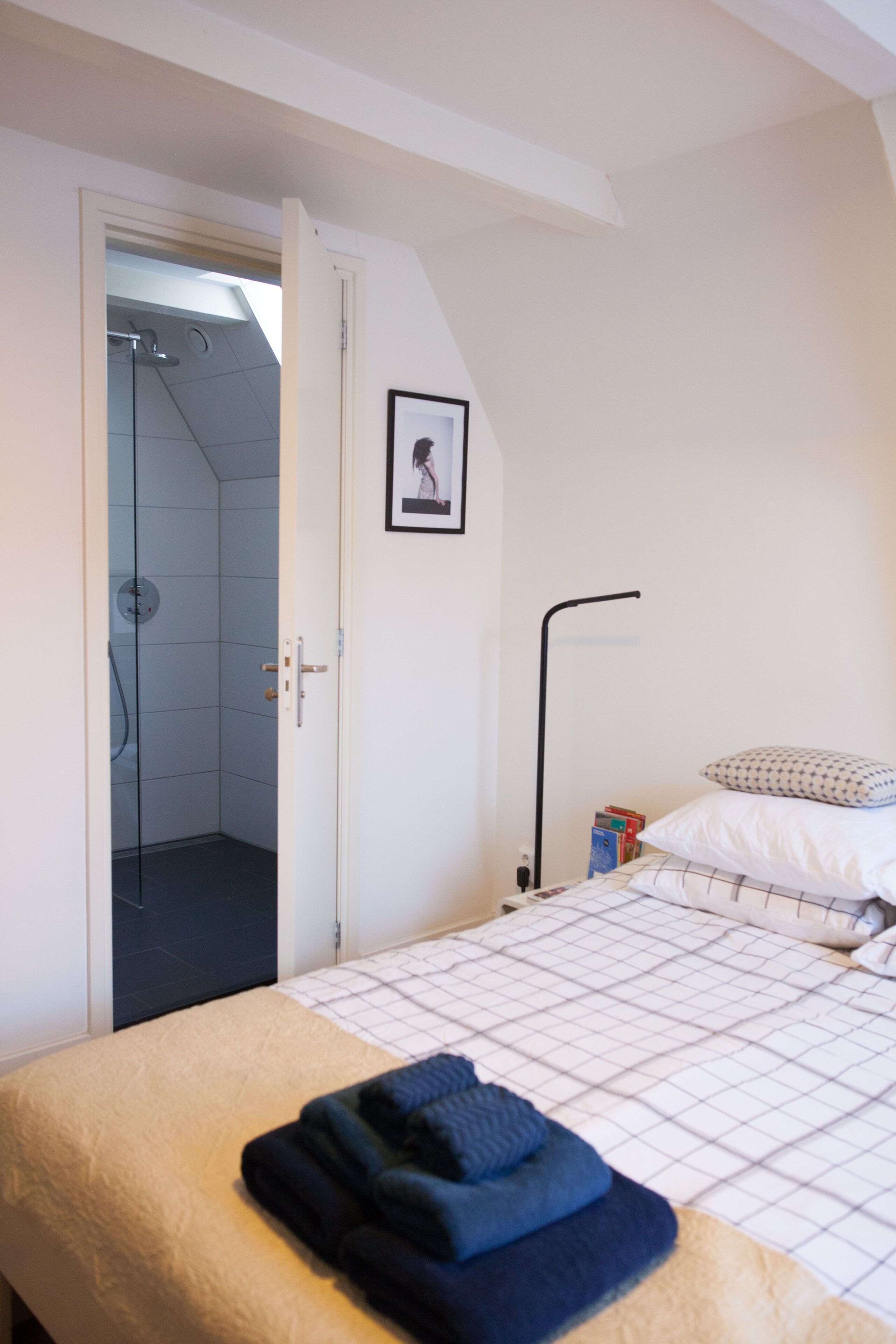 Et soveværelse med seng, sengebord, et lille bord ved siden af sengen, et billede i ramme på væggen, og en dør til et badeværelse med bruser.