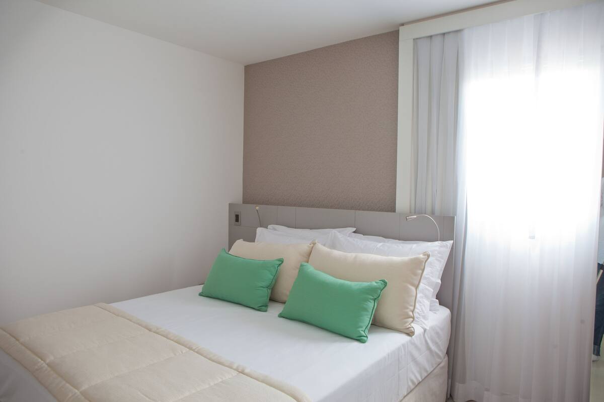 A neatly made bed with white and green pillows, a beige headboard, and a window with sheer curtains.