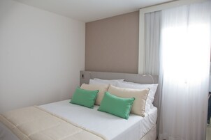 A neatly made bed with white and green pillows, a beige headboard, and a window with sheer curtains.