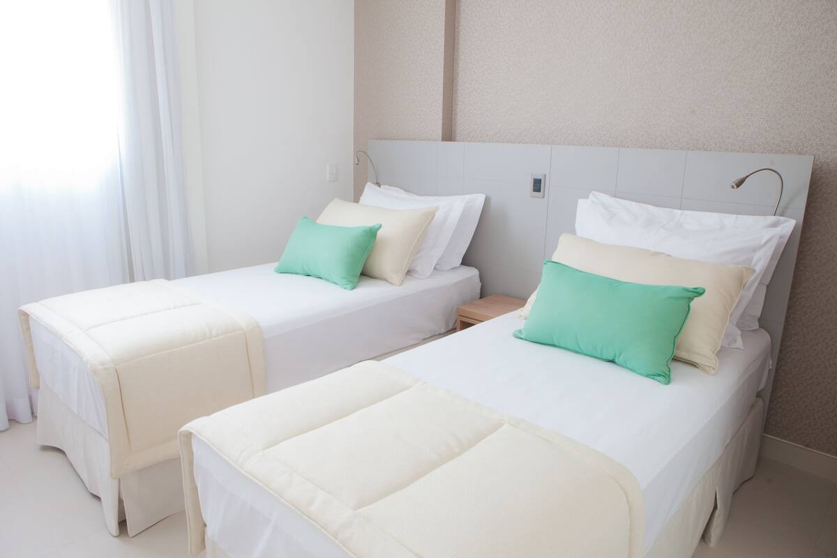 Two single beds with white bedding and teal pillows, a nightstand, and a wall-mounted lamp.