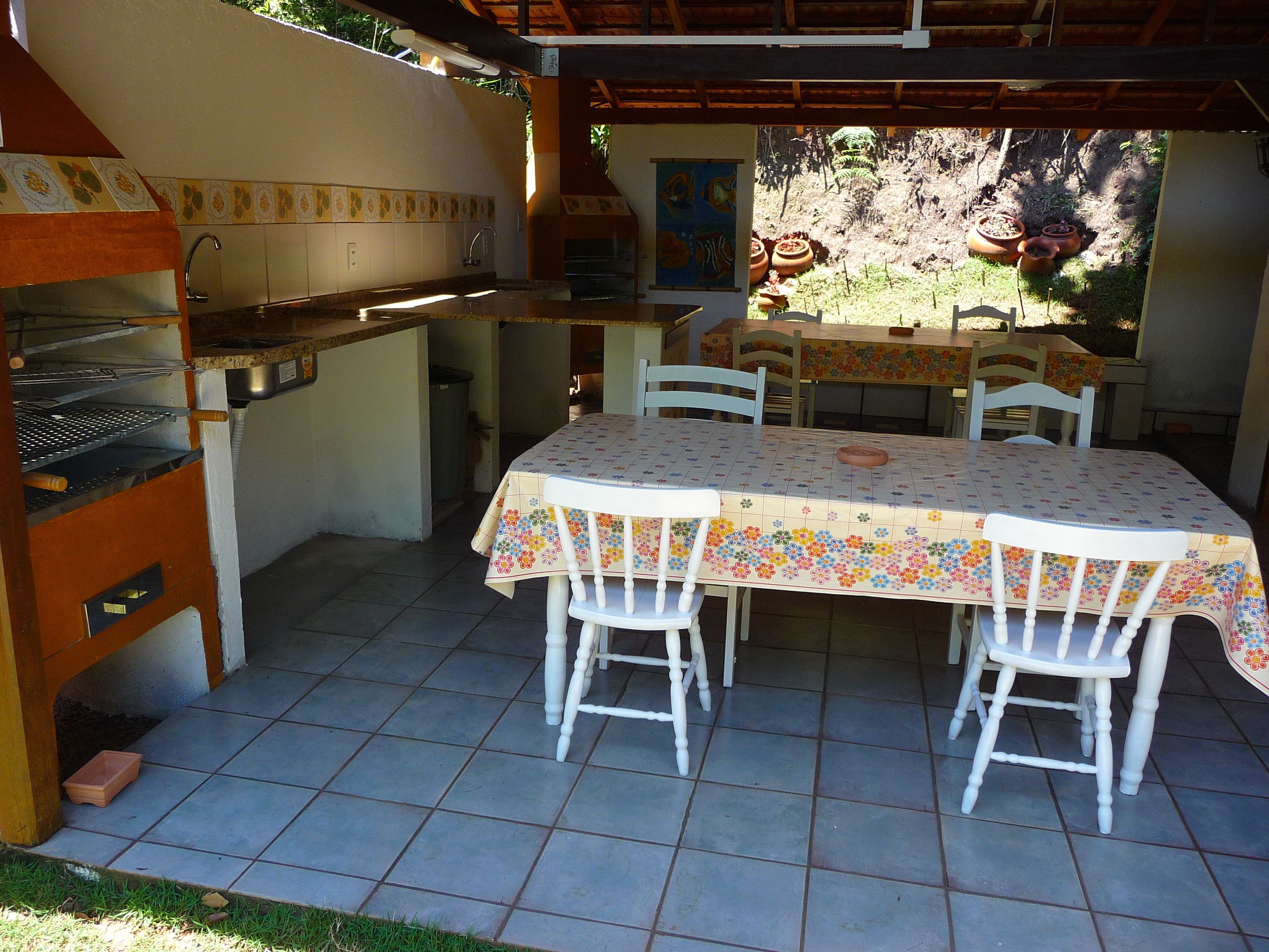 An outdoor patio with a tiled floor, a table with a floral tablecloth, white chairs, a barbecue grill, and a view of a garden with potted plants.