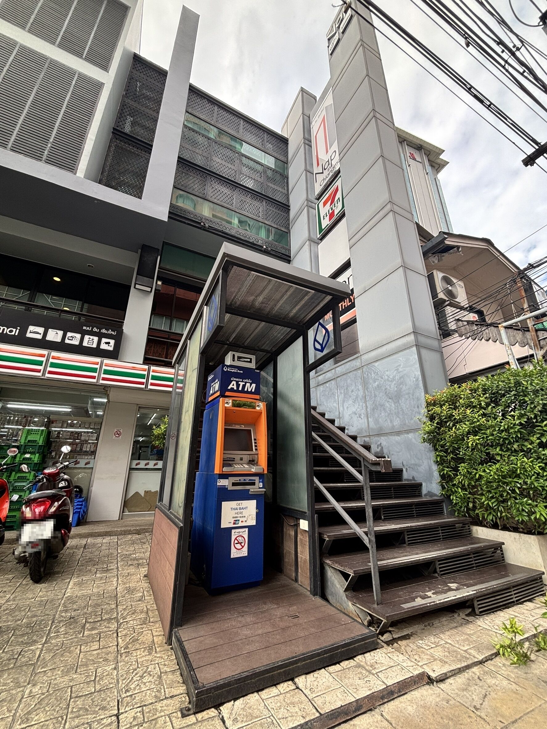 A 7-Eleven convenience store with an ATM, a staircase leading to a building, and a scooter parked nearby.