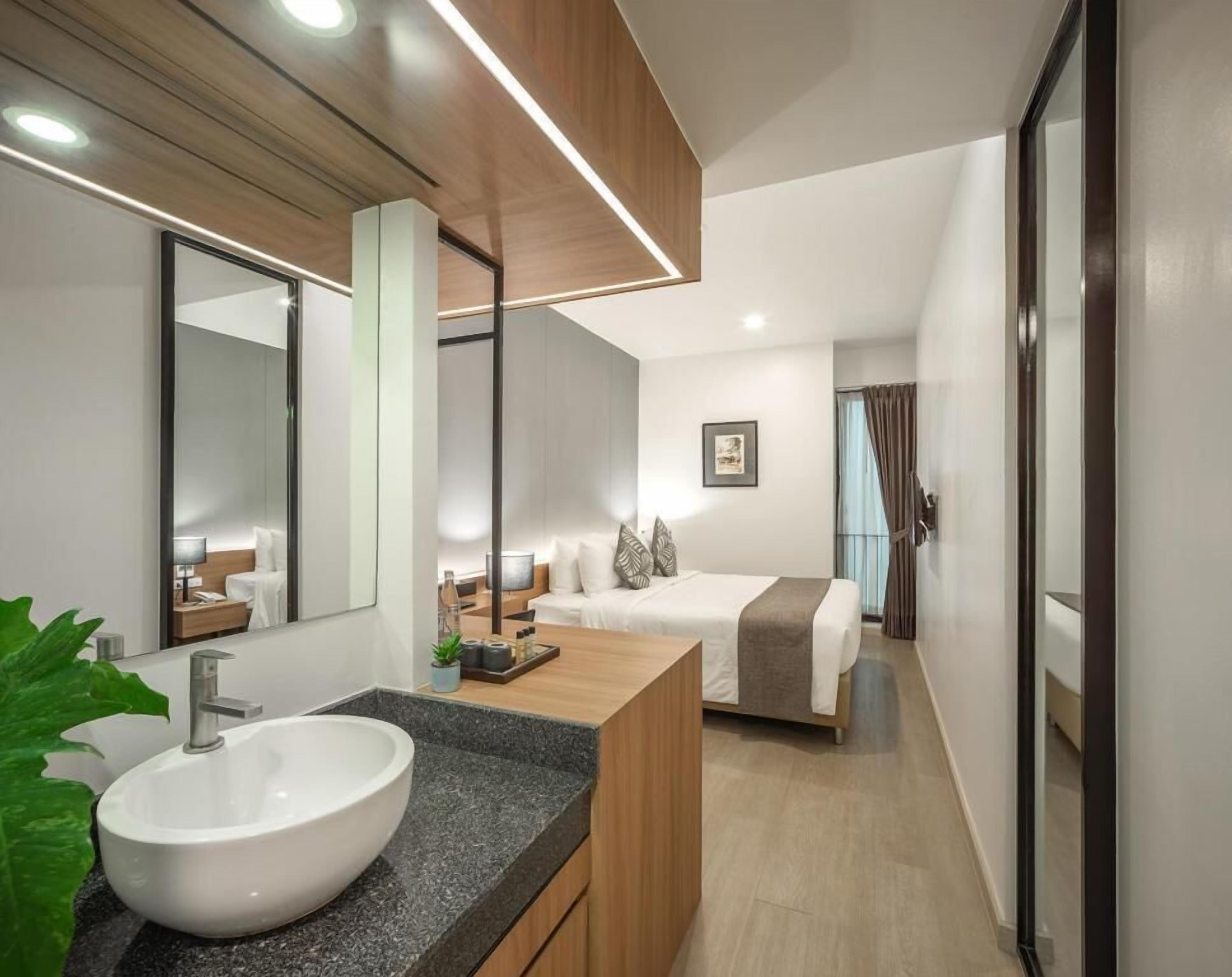 Une chambre d’hôtel moderne avec un grand miroir, un lavabo blanc, une coiffeuse en bois, un lit avec une literie blanche et une petite table de chevet.