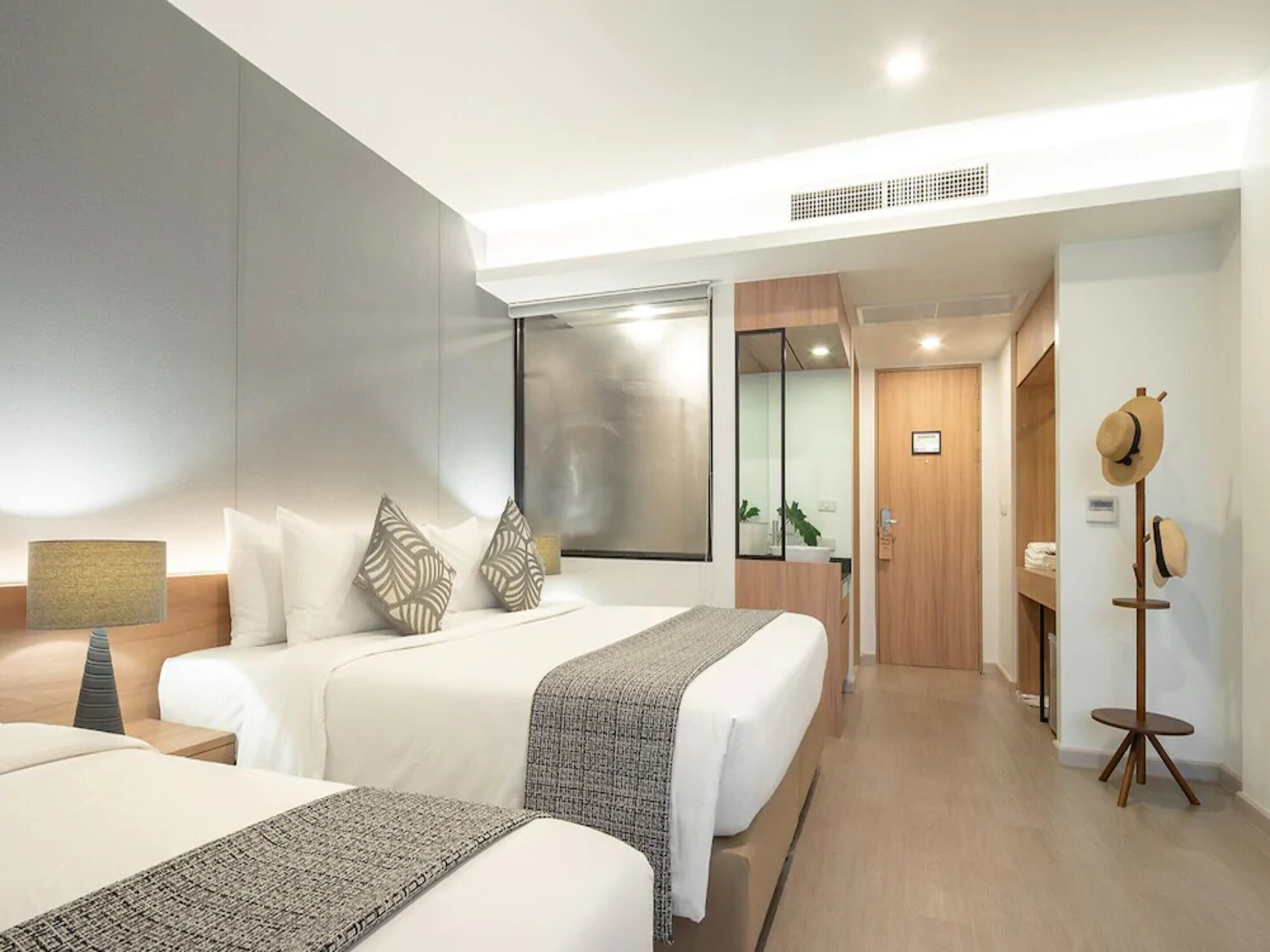 A modern hotel room with two beds, a large window, a mirror, a wooden door, and a coat rack.