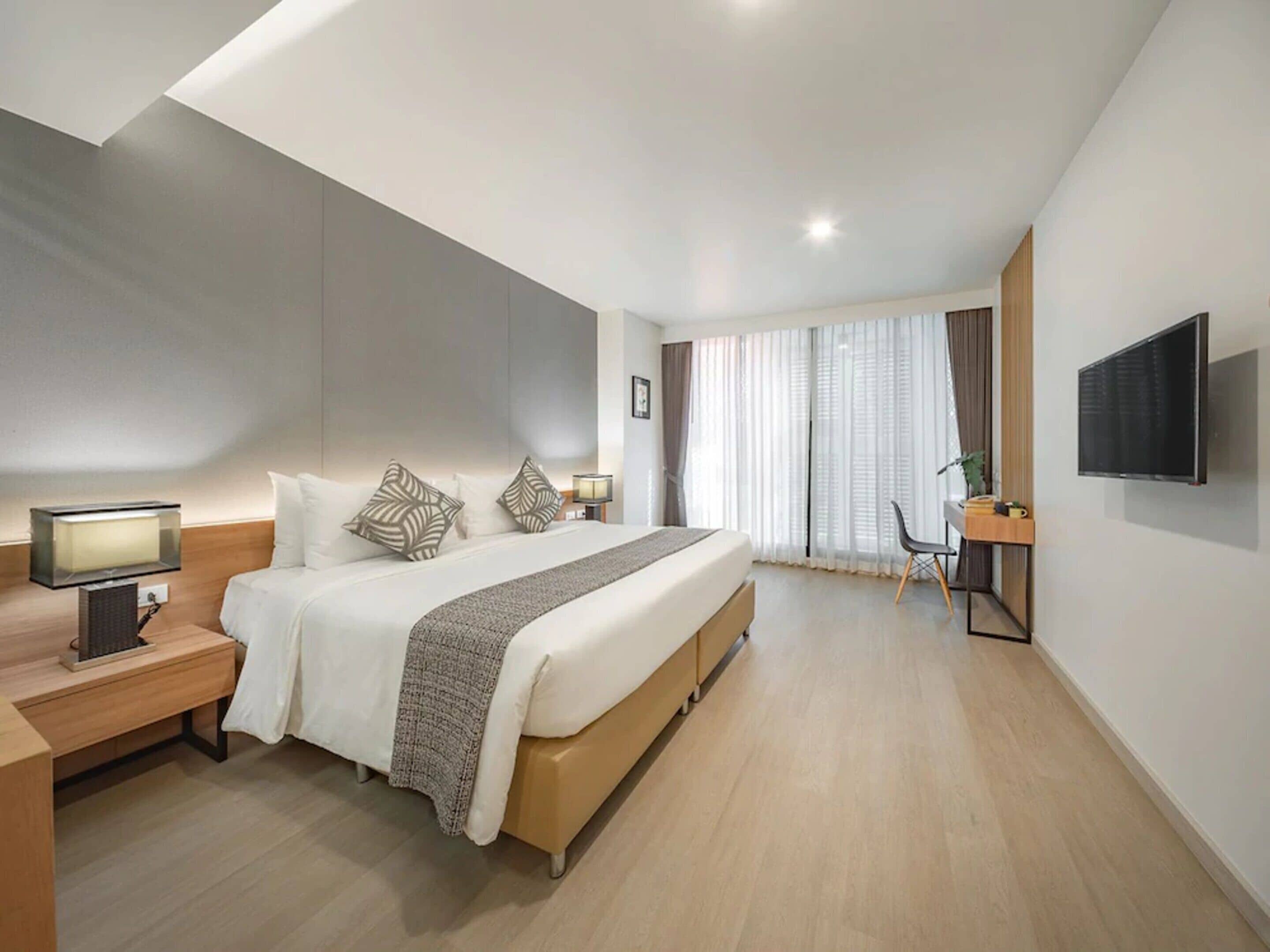 A modern hotel room with a large bed, bedside tables, a desk, a chair, and a TV.