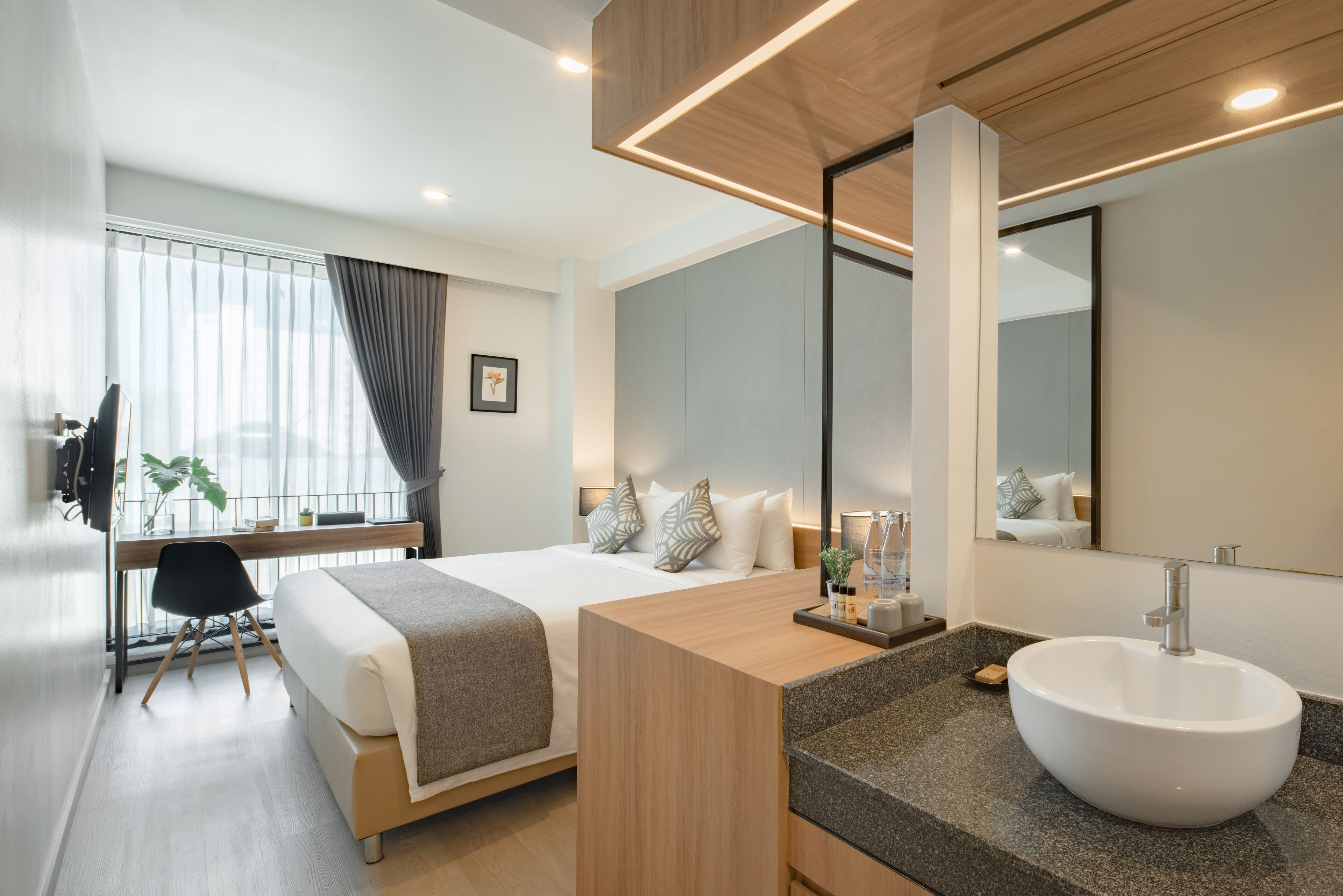 A modern hotel room with a large bed, a wooden desk, a large mirror, and a bathroom with a white sink.